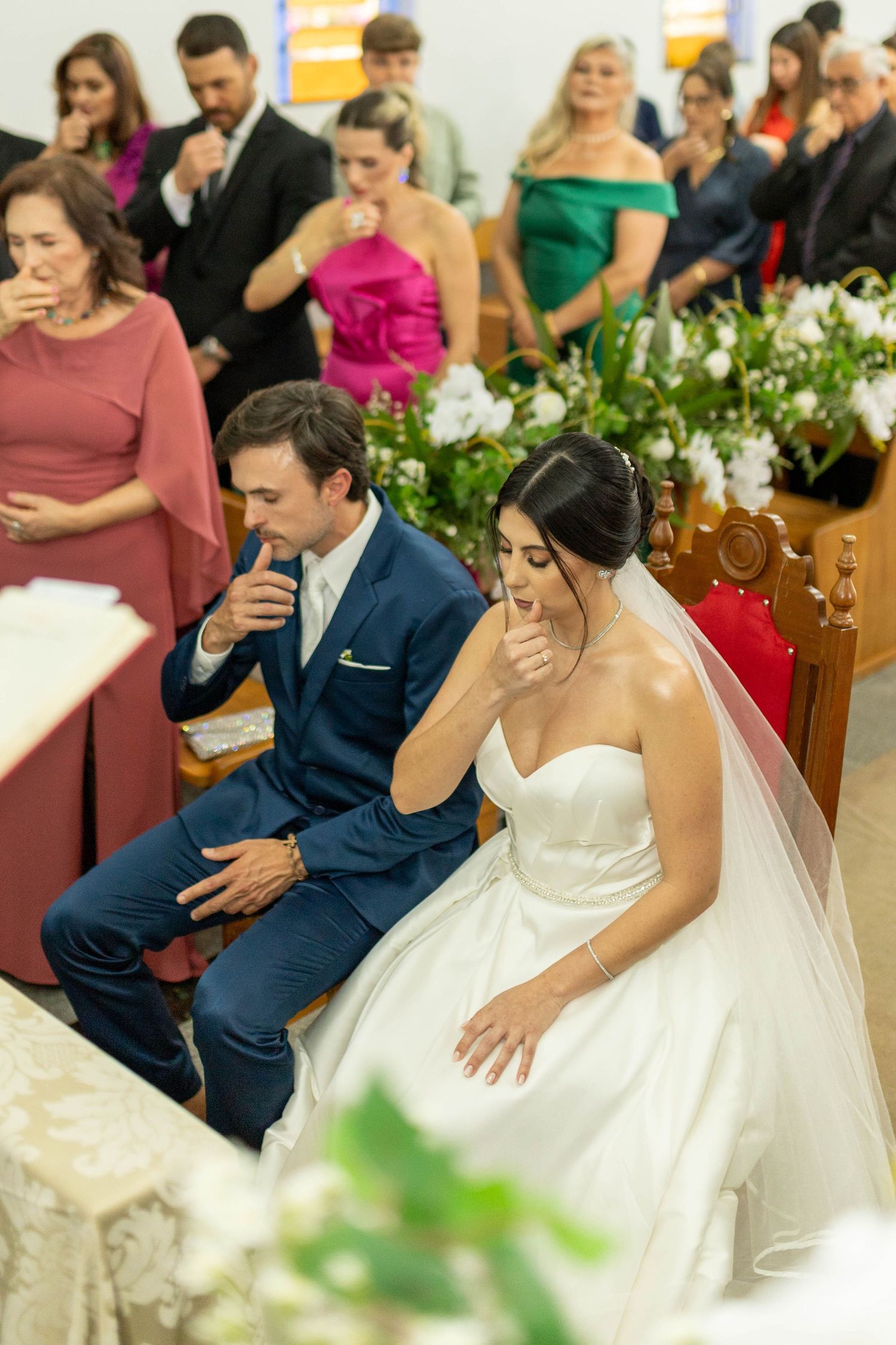 Bride in white gown and groom in blue suit kneel during wedding ceremony with guests and floral arrangements in background.