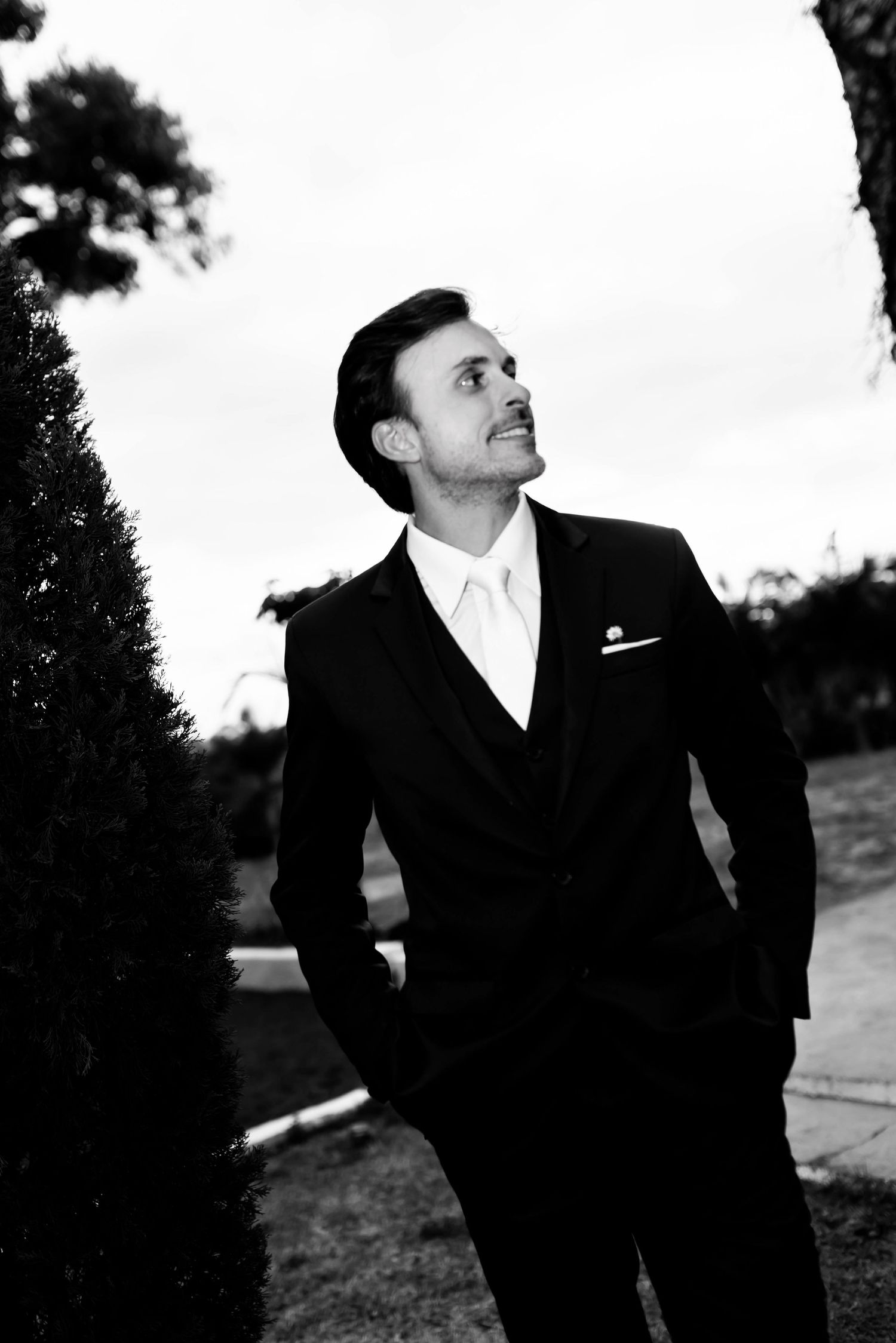 Black and white photo of an elegant man in a suit and tie posing outdoors among trees.