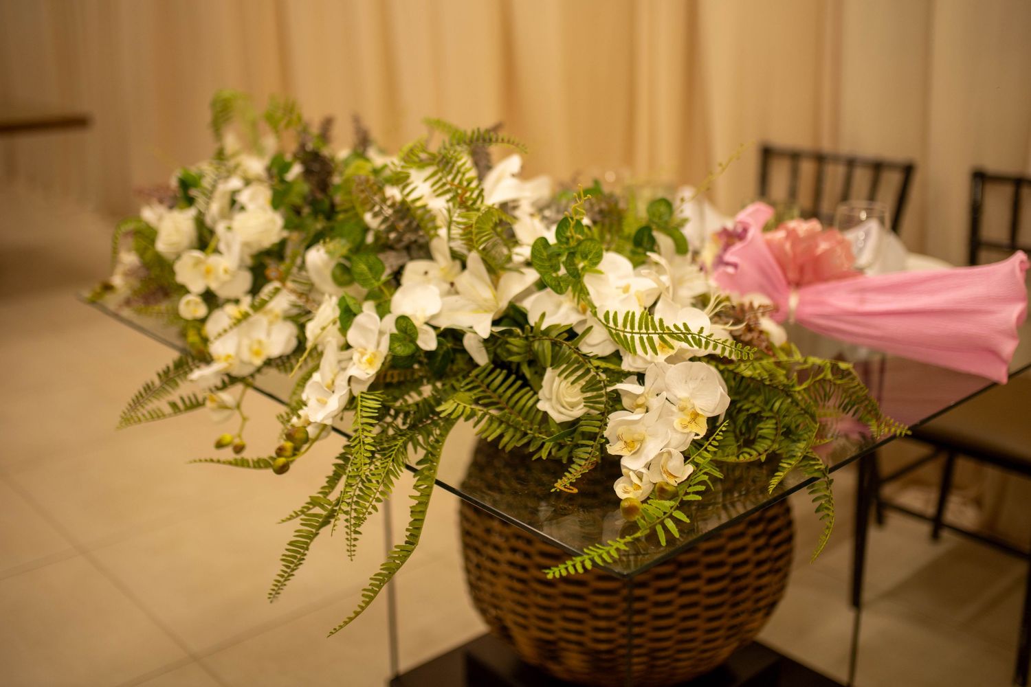 Elegant wicker basket centerpiece with white orchids and lush green ferns on a glass table at a formal event.