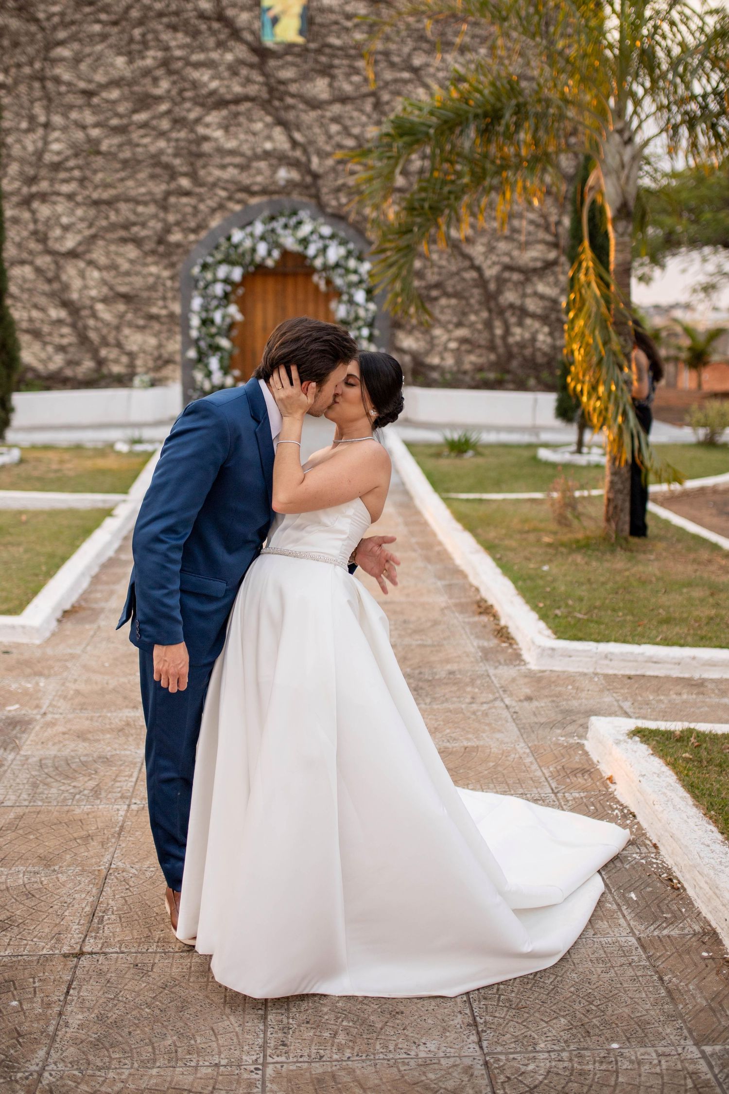 Bride and groom share a romantic kiss outside a stone church, bride in white ballgown, groom in navy suit.