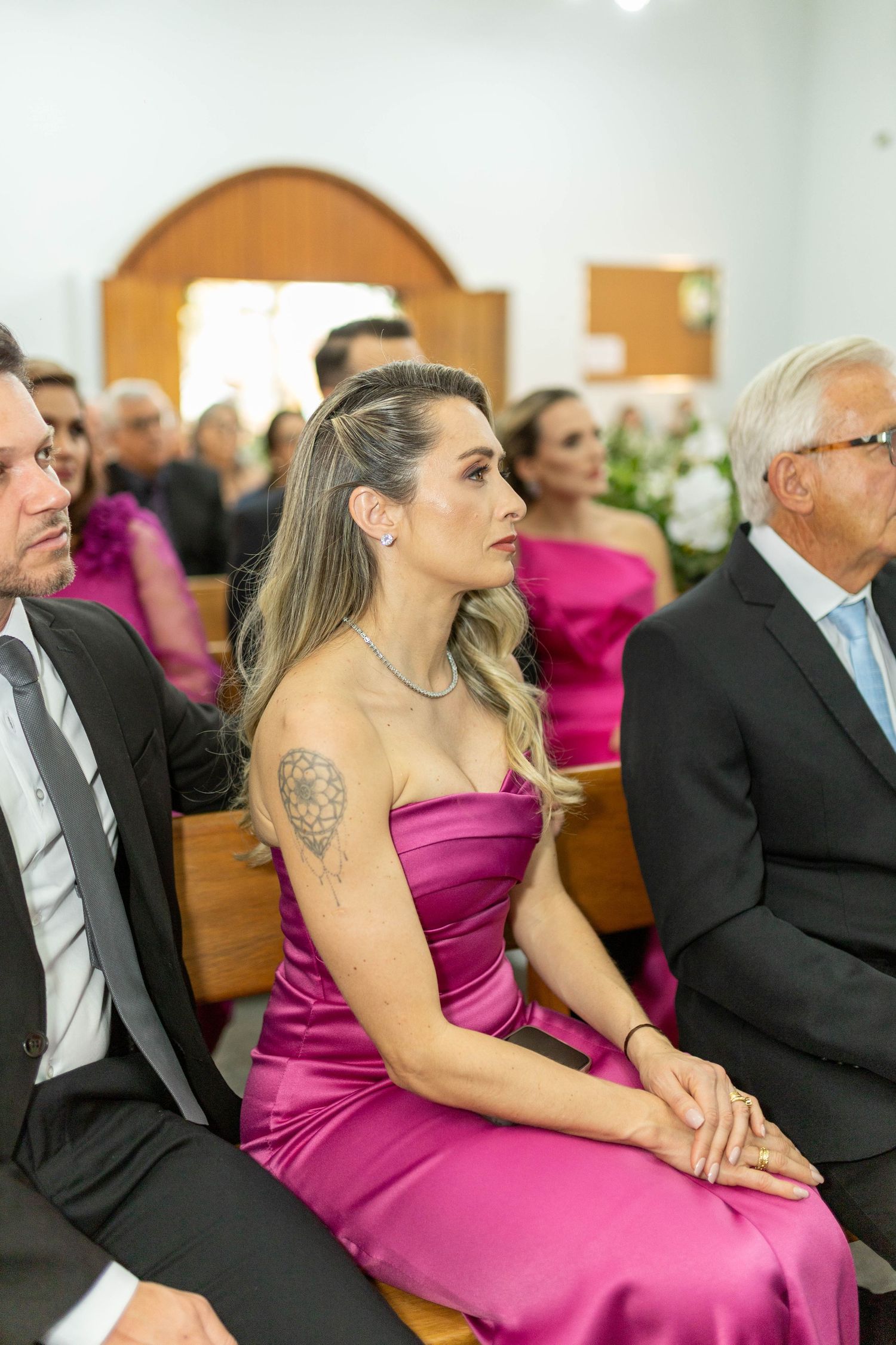Woman in magenta strapless satin gown with tattoo sits attentively at a wedding ceremony in a church.