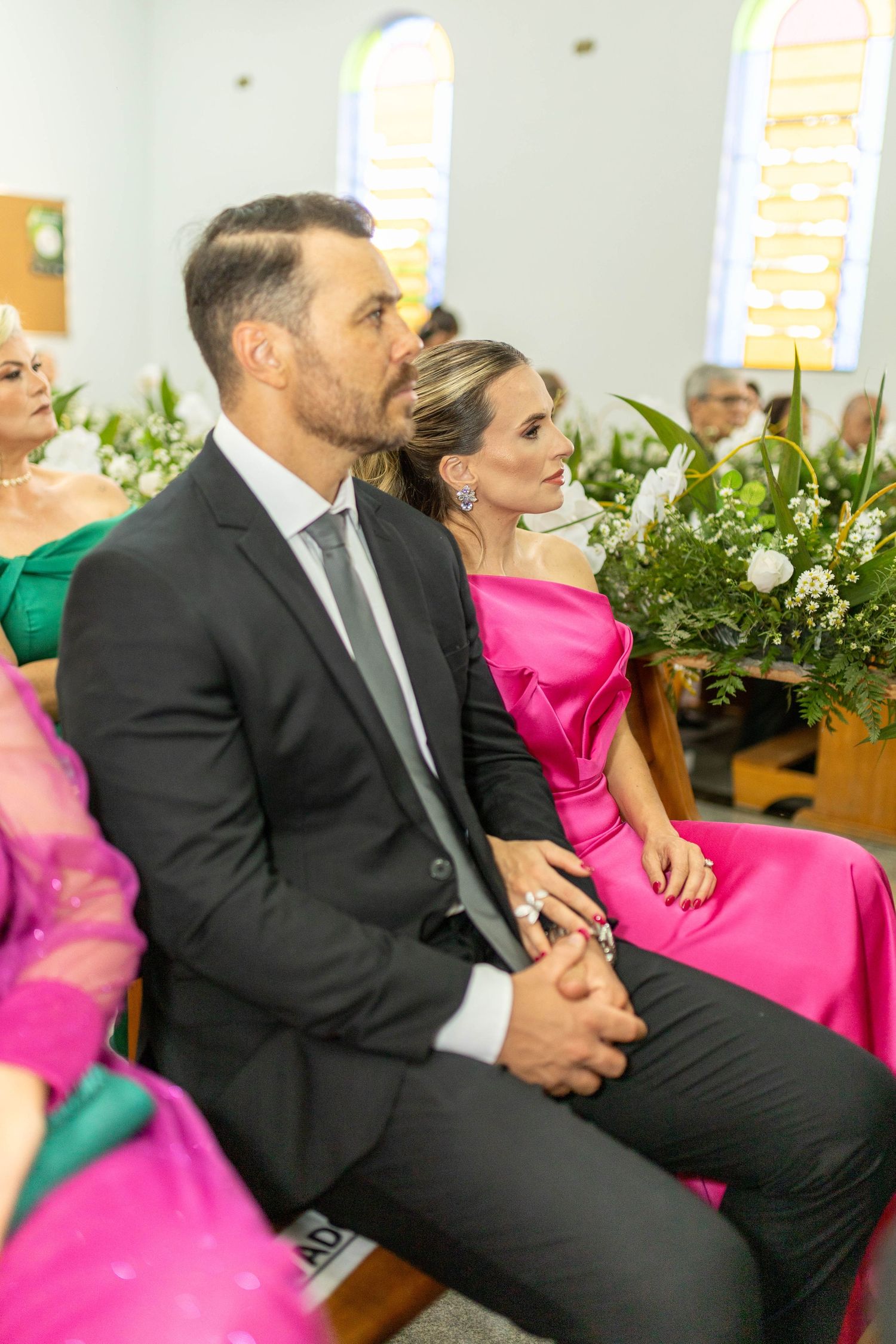 Elegant couple in formal attire sitting in church pews at a wedding ceremony, man in suit, woman in pink dress.