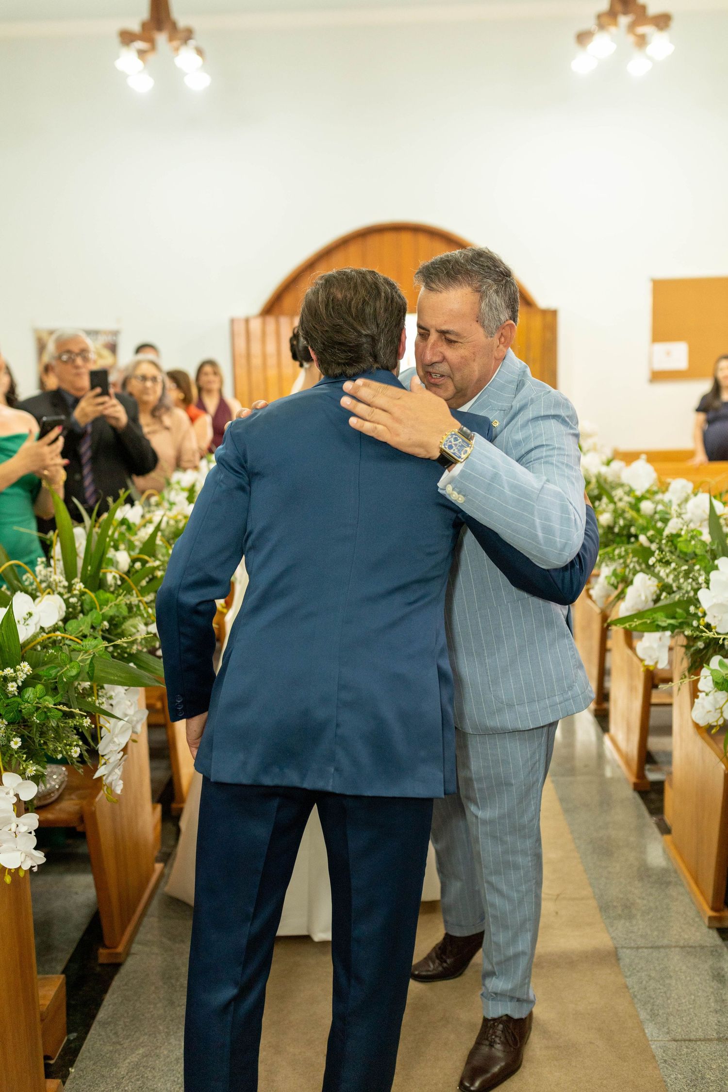 Two men in suits embrace warmly at a church wedding ceremony, surrounded by floral decorations and guests.