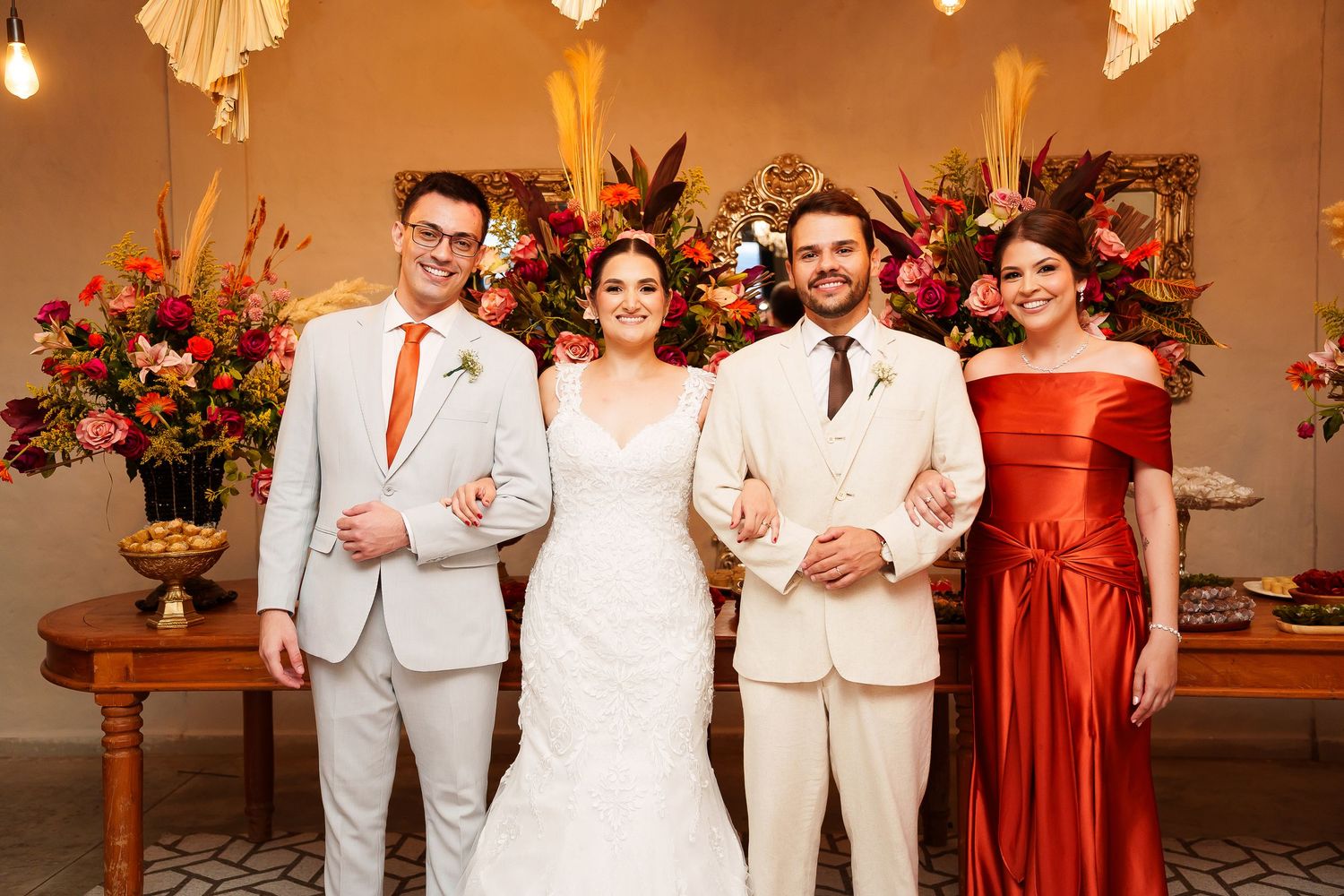Bride and groom pose with two guests at a wedding ceremony, surrounded by vibrant floral arrangements.