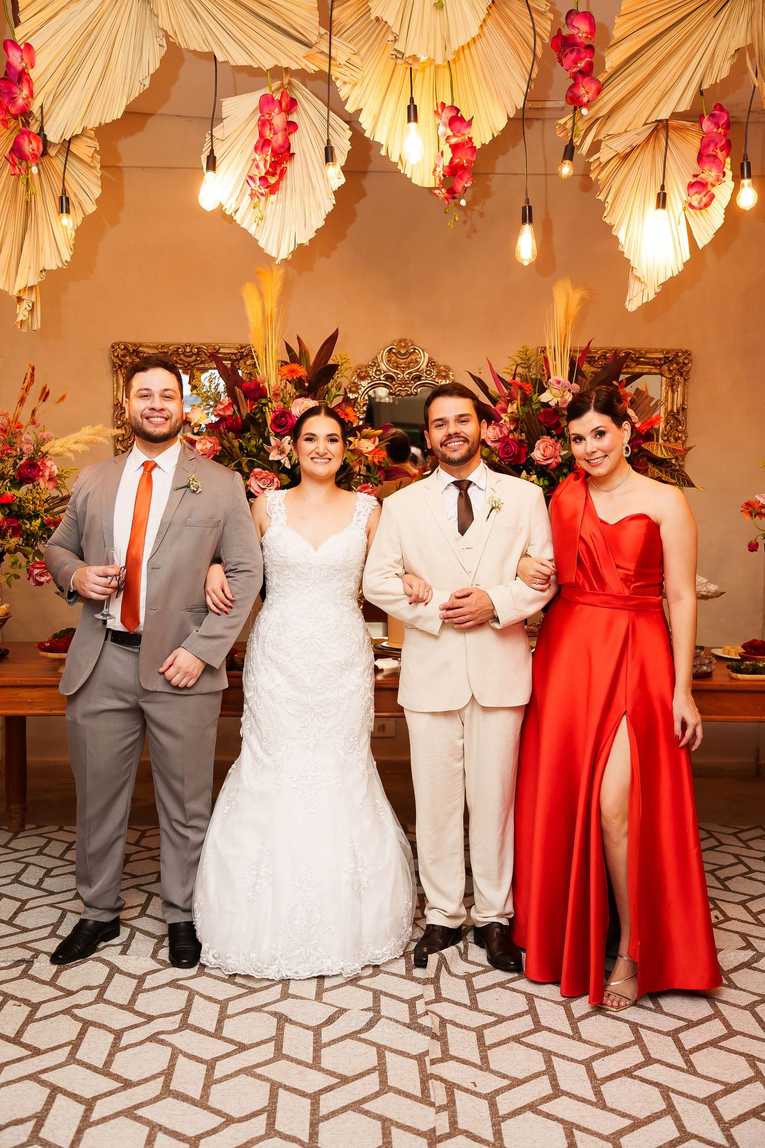 Two couples pose at a wedding reception, bride in white lace gown, groom in cream suit, guests in gray suit and red dress.