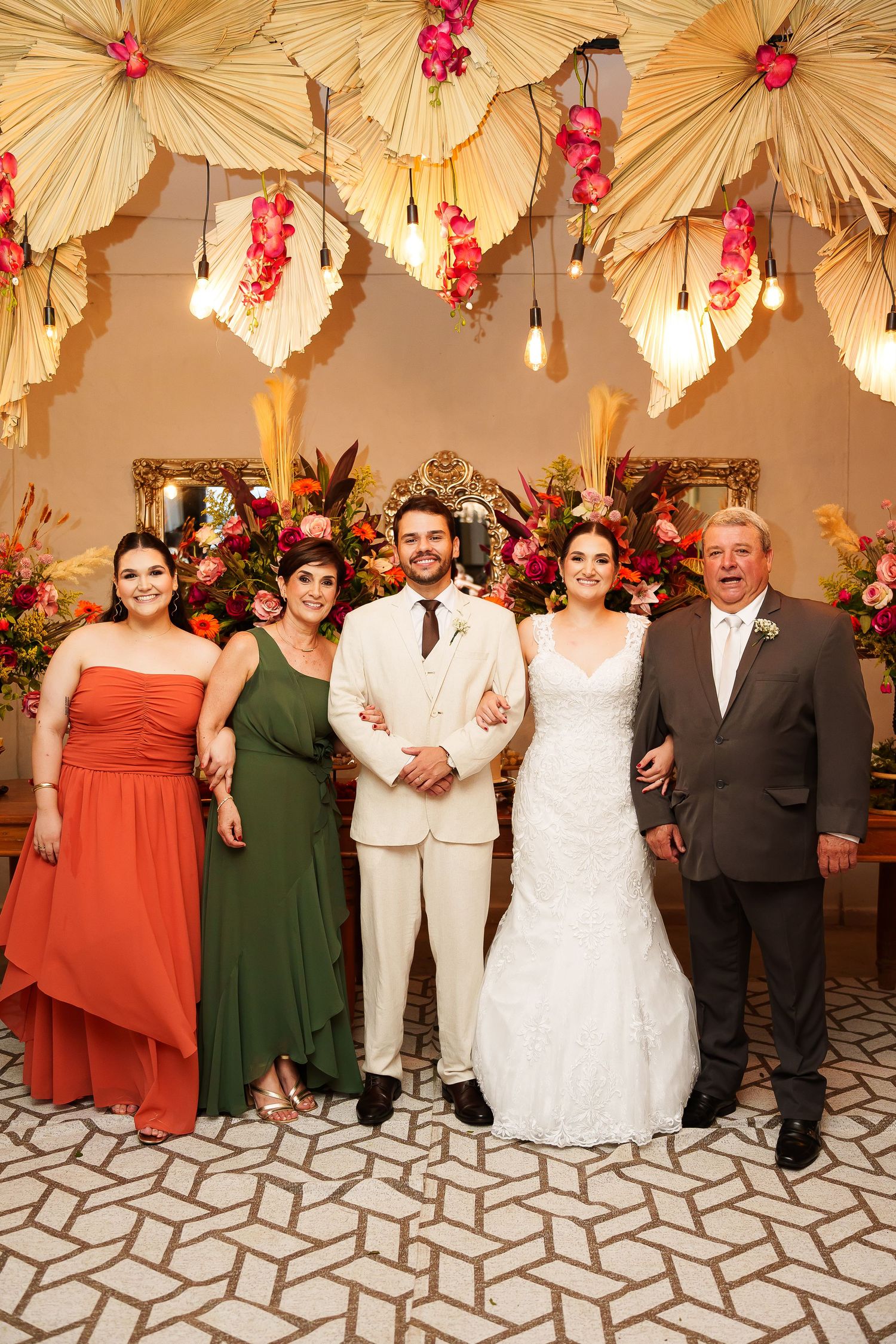 Wedding party of five posing elegantly under floral decorations, bride in white gown flanked by family in formal attire.