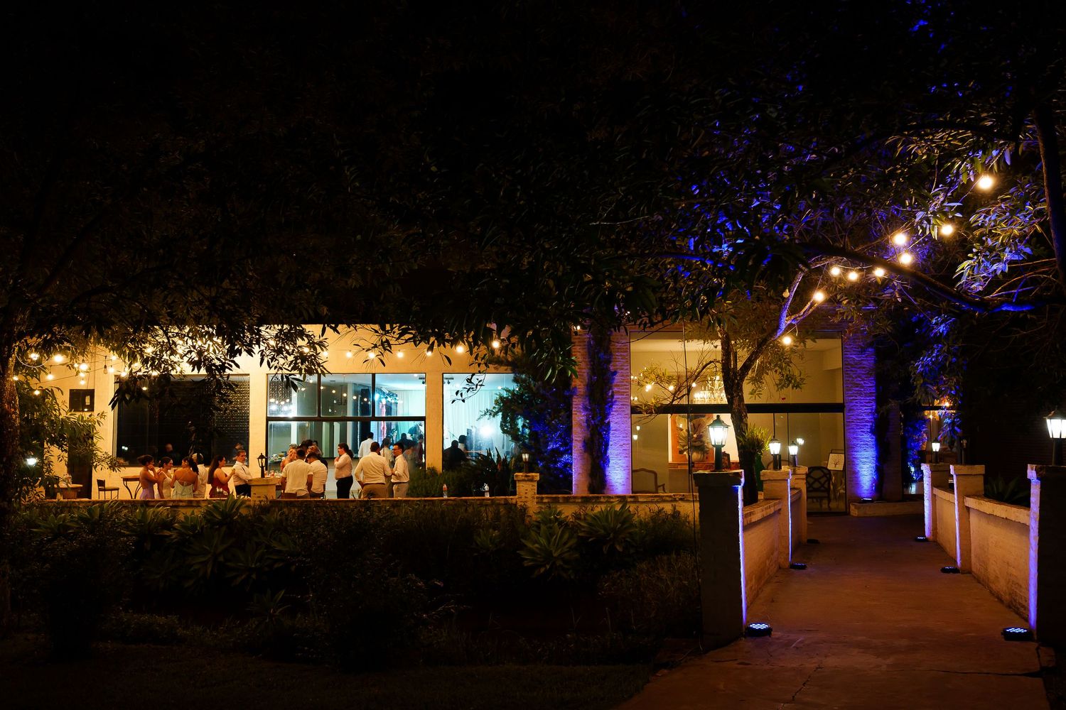 Outdoor restaurant patio glowing with string lights and blue uplighting at night, surrounded by trees.