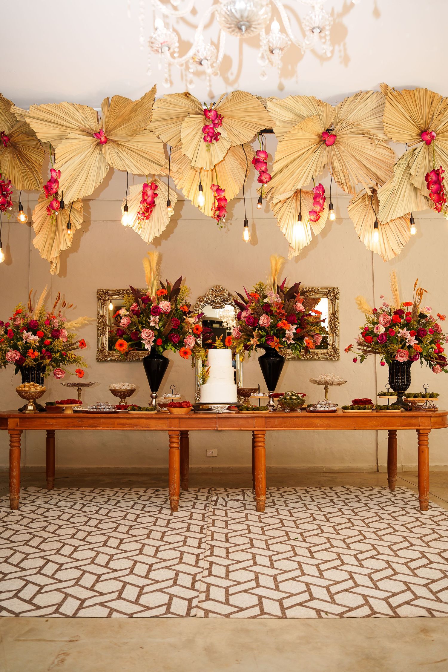 Elegant wedding dessert table with white cake, tropical floral arrangements, and decorative palm leaf backdrop with hanging lights.