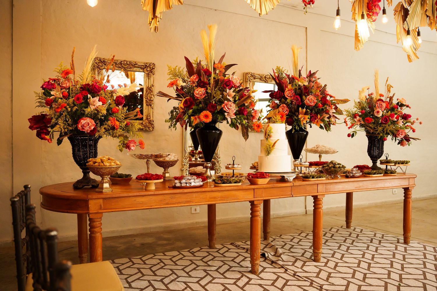 Elegant fall wedding reception table with large floral arrangements in dark urns, wedding cake, and autumn decor.
