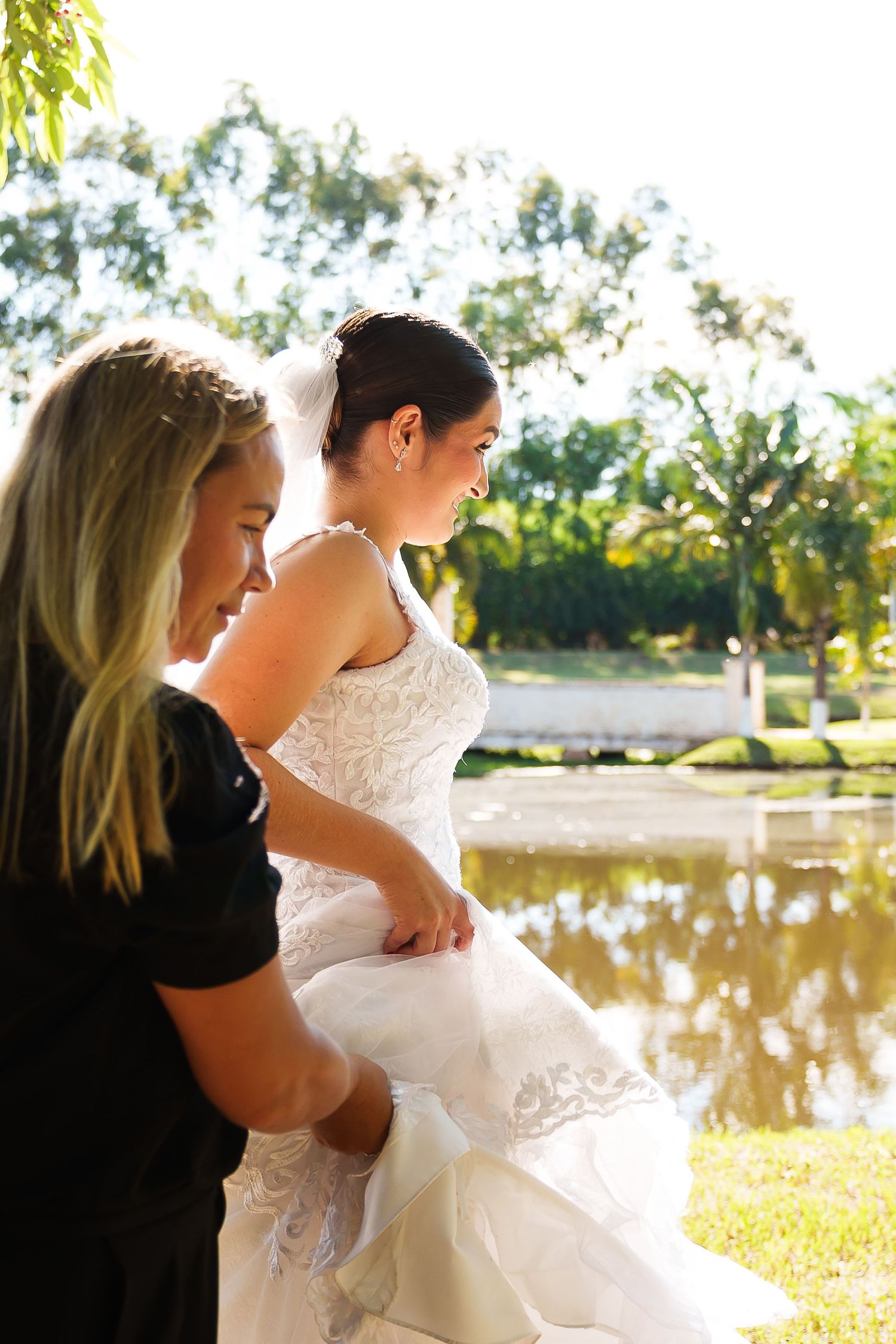 A bride in a white lace gown stands by a pond as a blonde woman in black adjusts her dress outdoors.