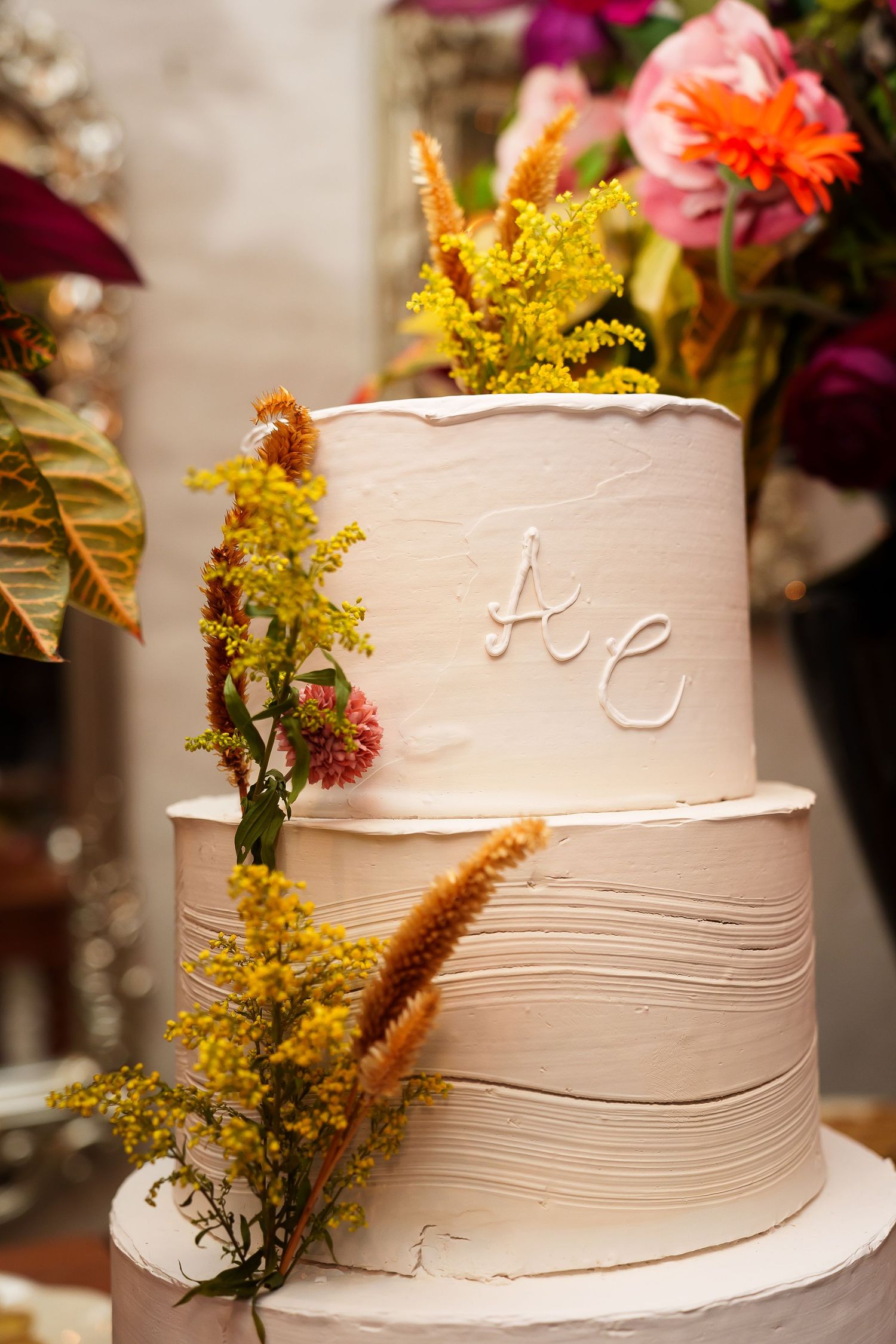 Two-tier white textured wedding cake with yellow wildflowers and monogram initials, surrounded by colorful floral arrangements.