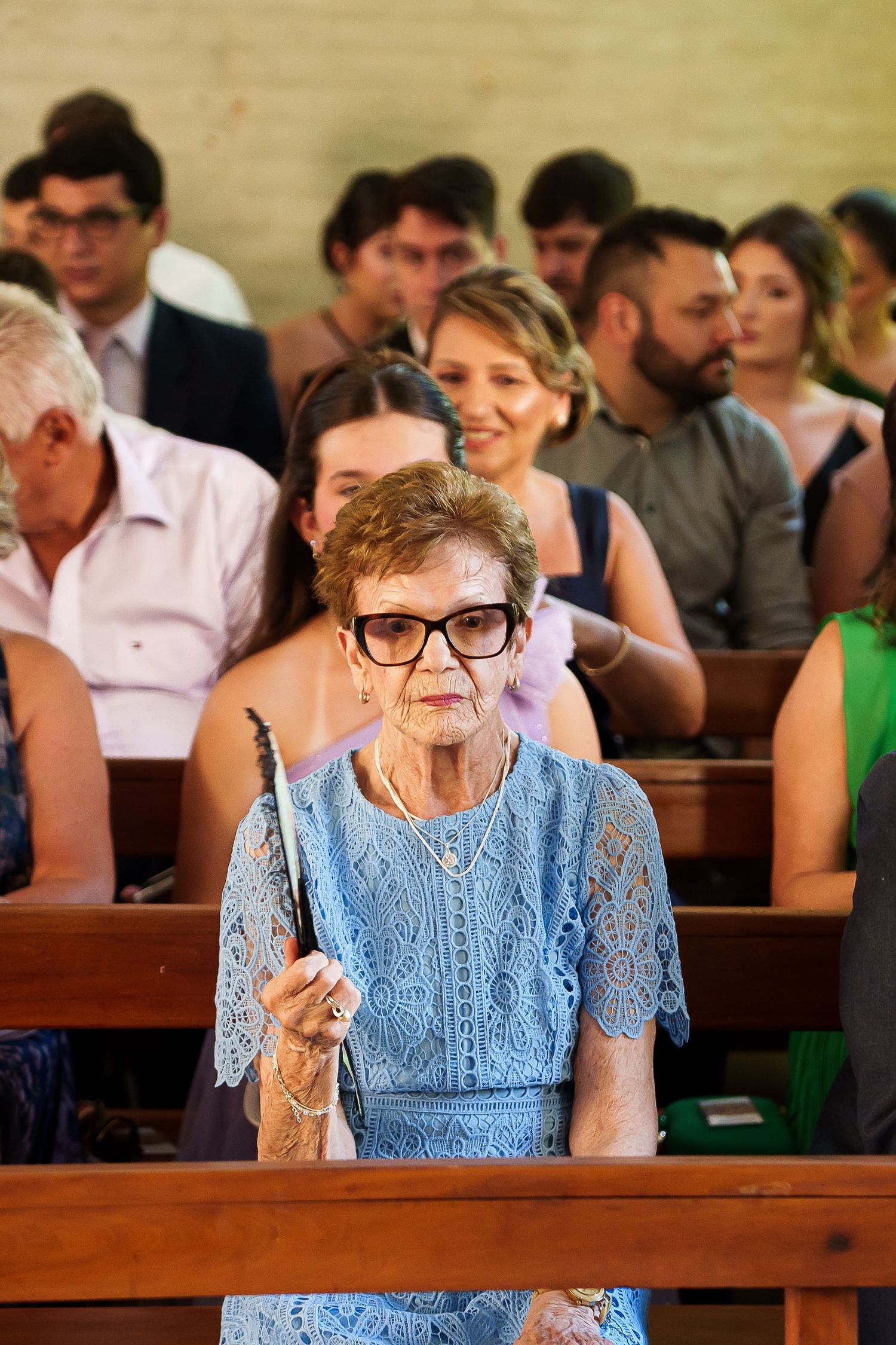 Elderly woman in blue lace dress holding a fan sits in church pew during a wedding ceremony.