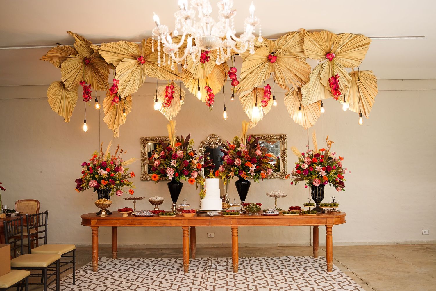 Elegant wedding dessert table with tropical palm leaf decor, floral arrangements, and chandelier in a bright reception hall.