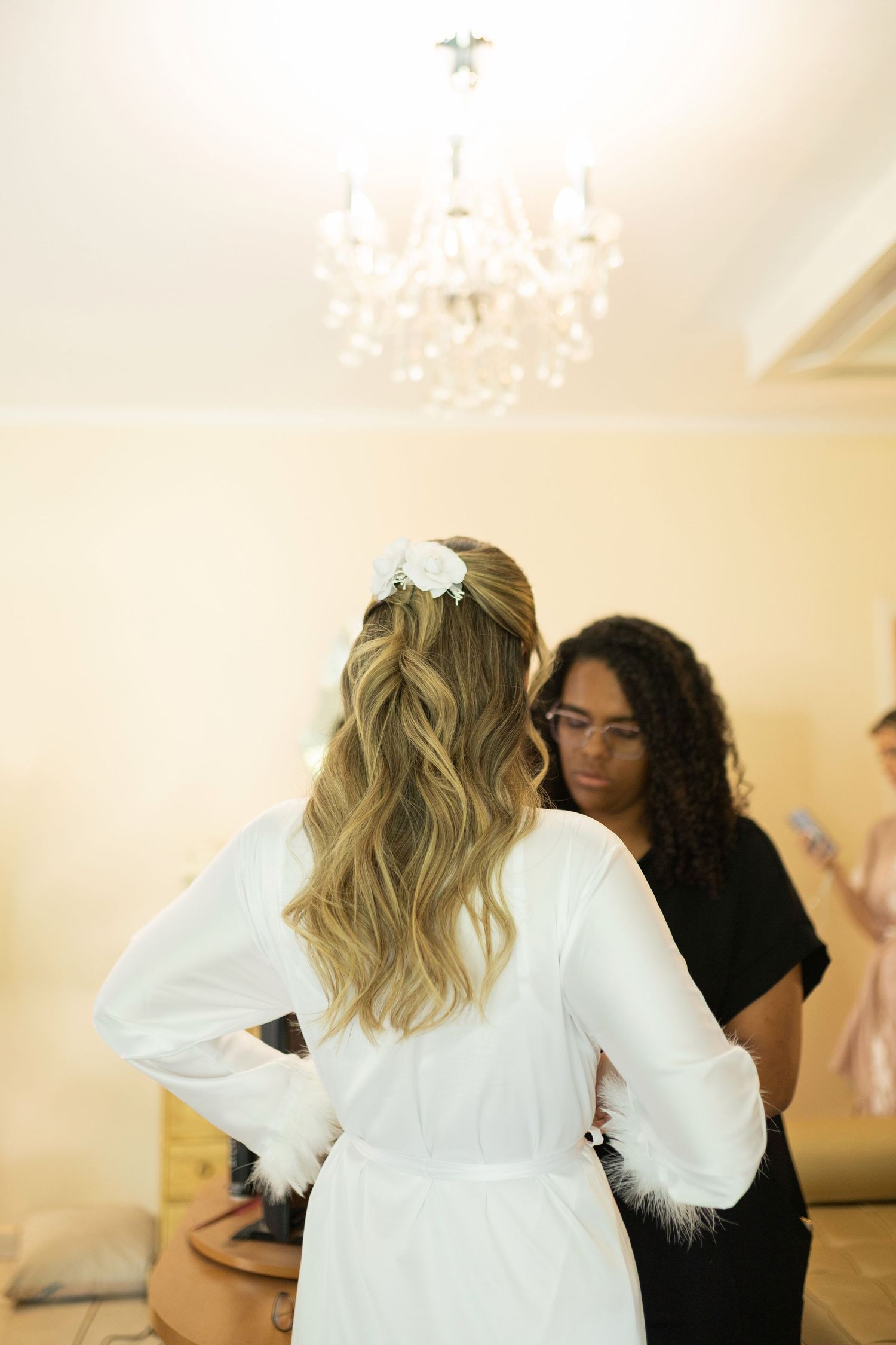 Bride in white robe with floral hair accessory getting ready, assisted by stylist under crystal chandelier.