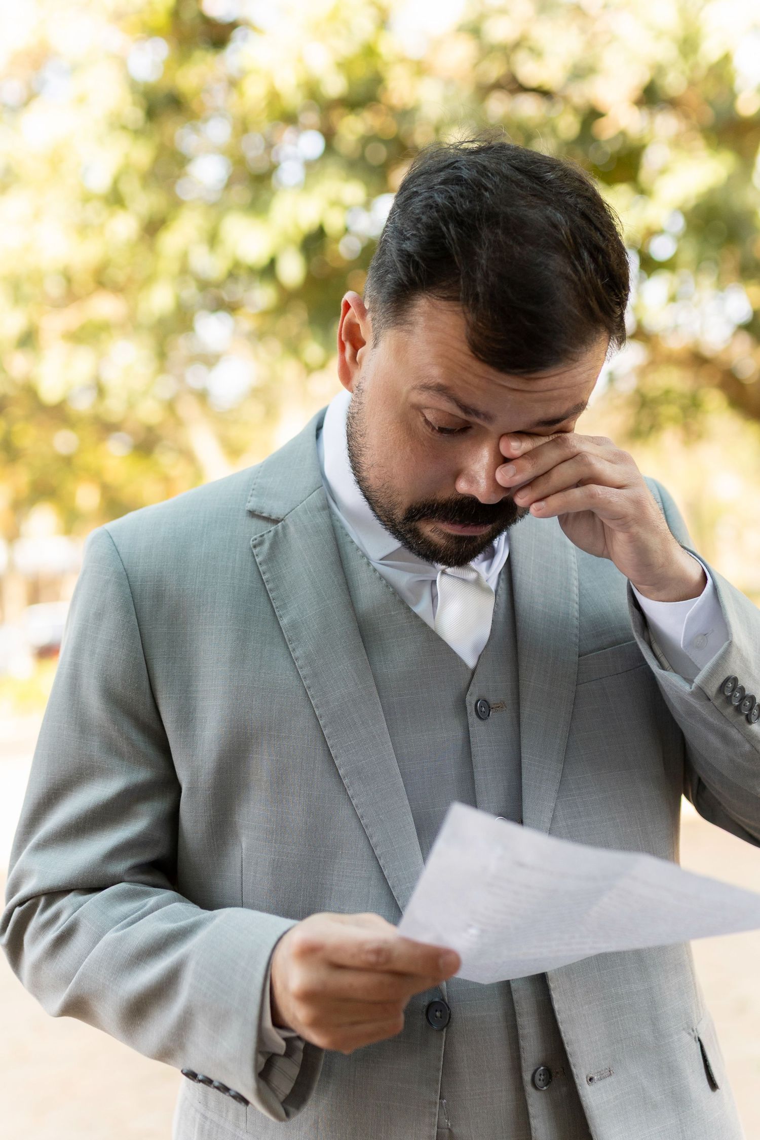Emotional bearded man in gray suit reading a letter outdoors, pinching his nose in distress.