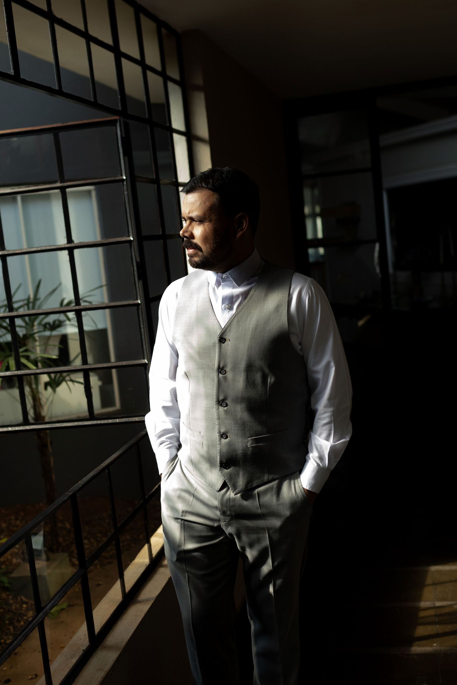 A well-dressed man in a white vest and shirt stands thoughtfully by a window in dramatic light.