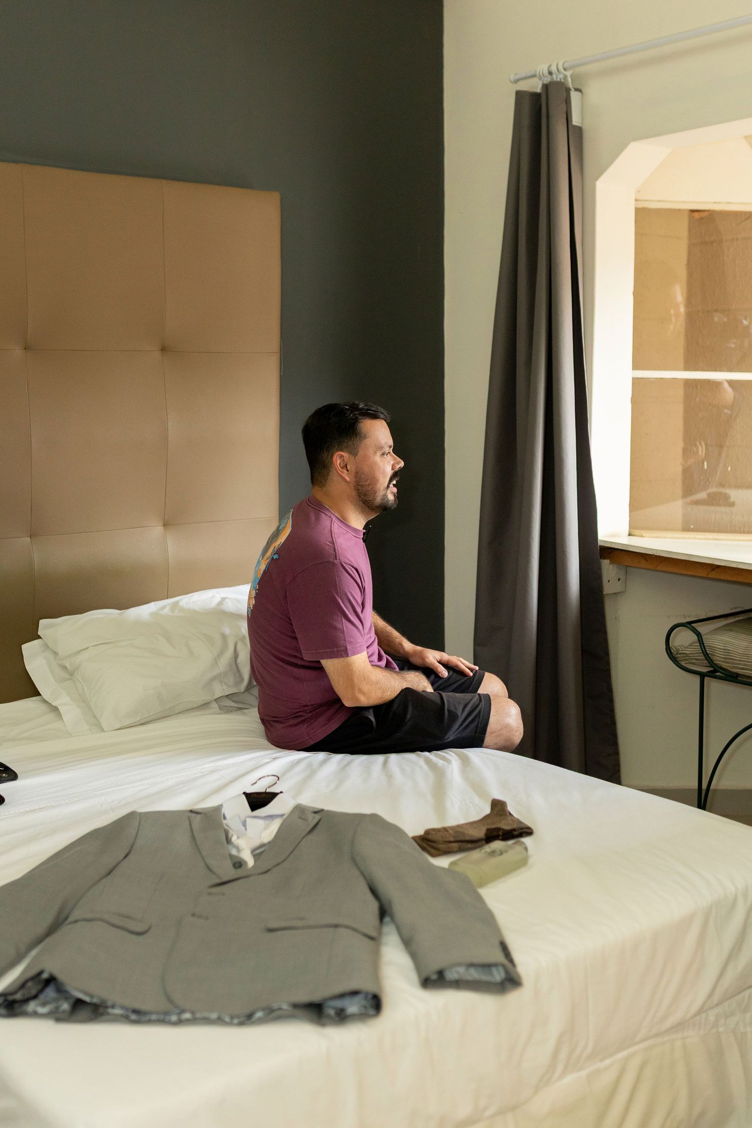 Man in purple shirt sitting on white bed beside a gray suit jacket and brown shoes in a modern bedroom.