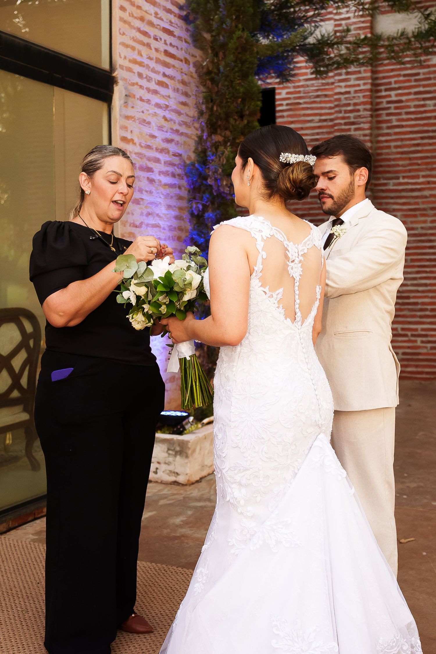Wedding coordinator in black dress assists bride in white lace gown and groom in tan suit with bouquet outdoors.