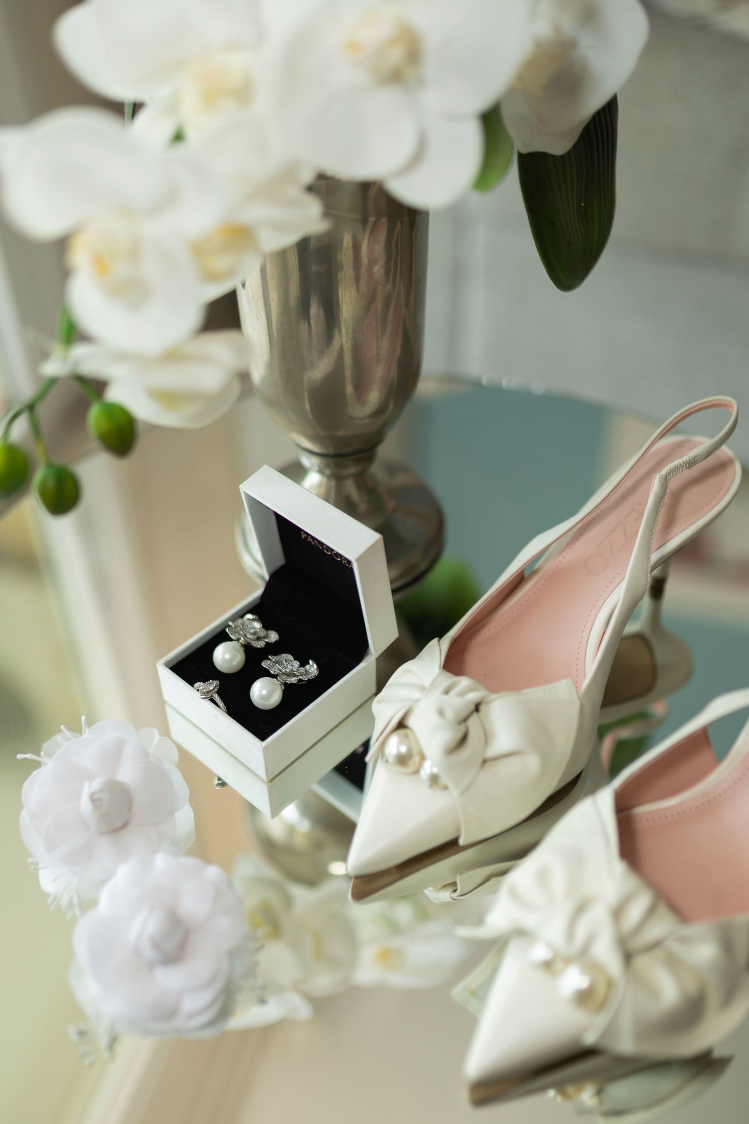 Elegant bridal accessories featuring white satin heels, pearl earrings in a gift box, and white orchids on a glass surface.