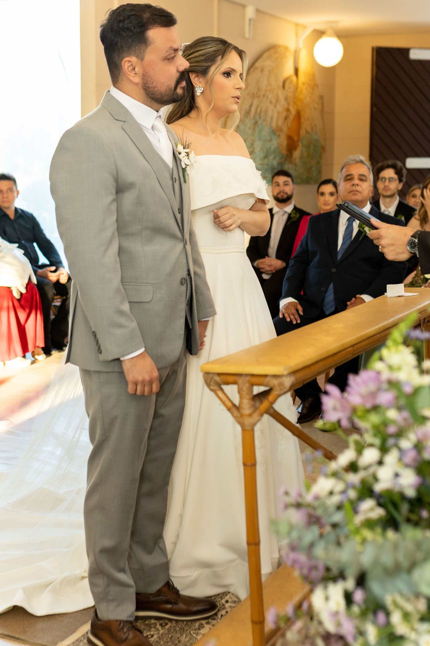 A bride and groom stand at the altar during their wedding ceremony, surrounded by guests in a decorated church.
