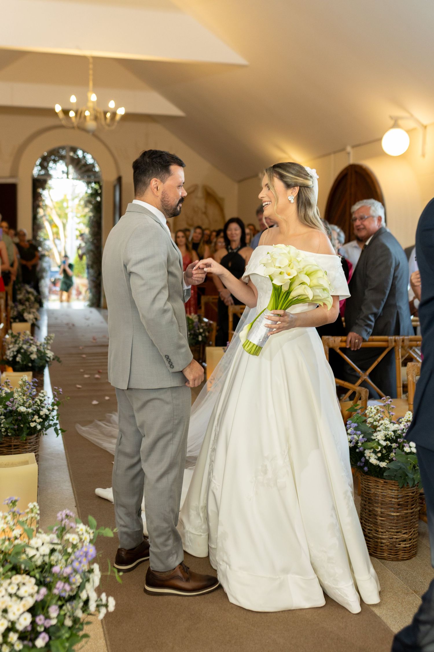 Bride and groom exchange rings at wedding ceremony, bride holding white calla lily bouquet in elegant off-shoulder gown.