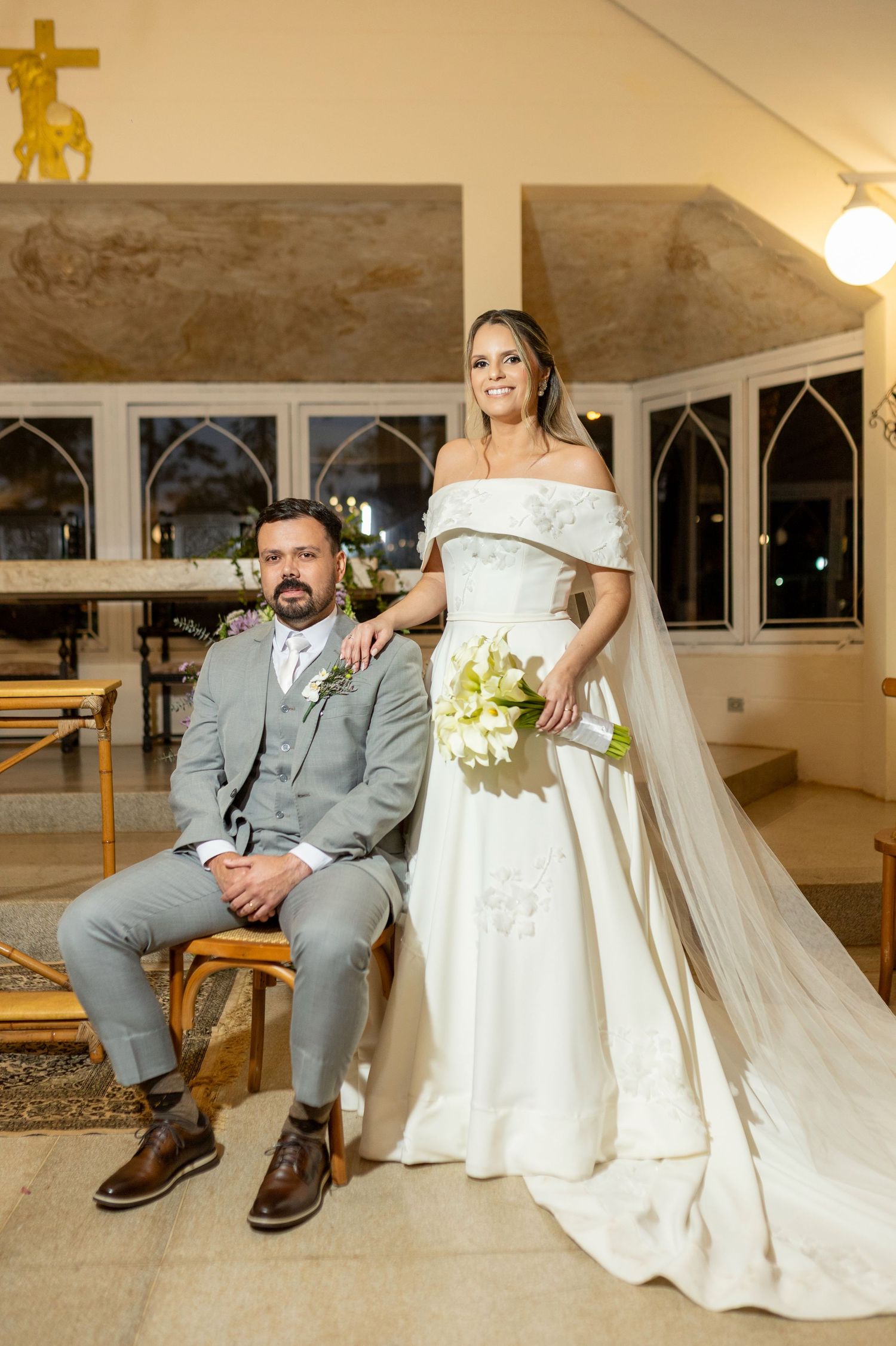 Bride and groom posing in a church, bride in off-shoulder white gown with bouquet, groom in gray suit.