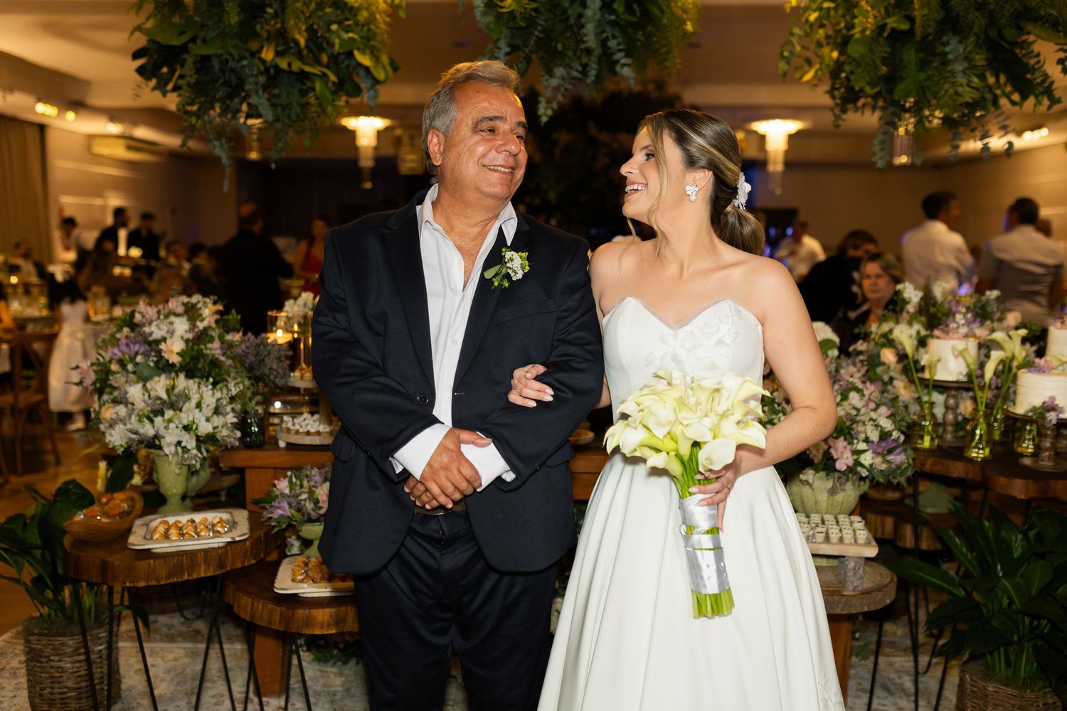 Bride in white gown holding bouquet stands with suited man at elegant floral wedding reception.