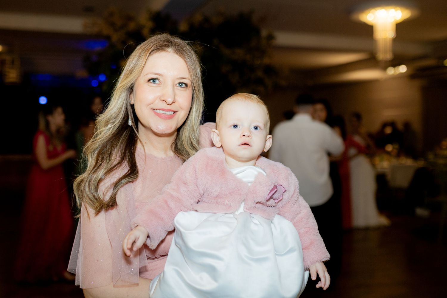 Woman in pink dress holding a baby in white outfit at an elegant indoor event.