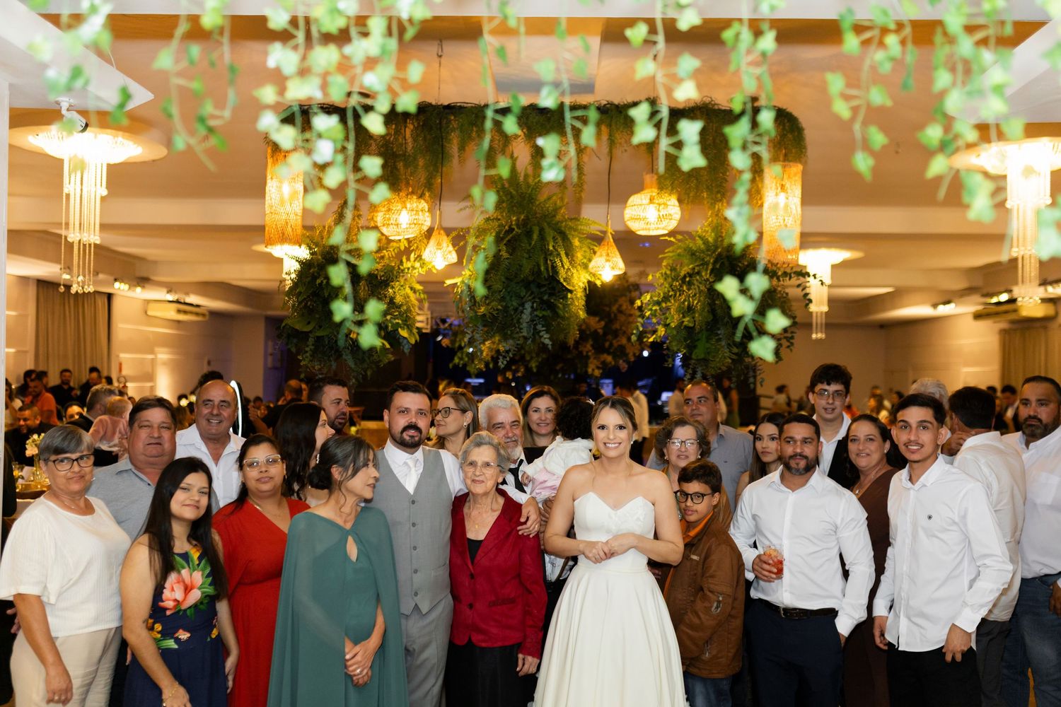 Bride in white gown poses with wedding guests in elegantly decorated reception hall with greenery ceiling decor.
