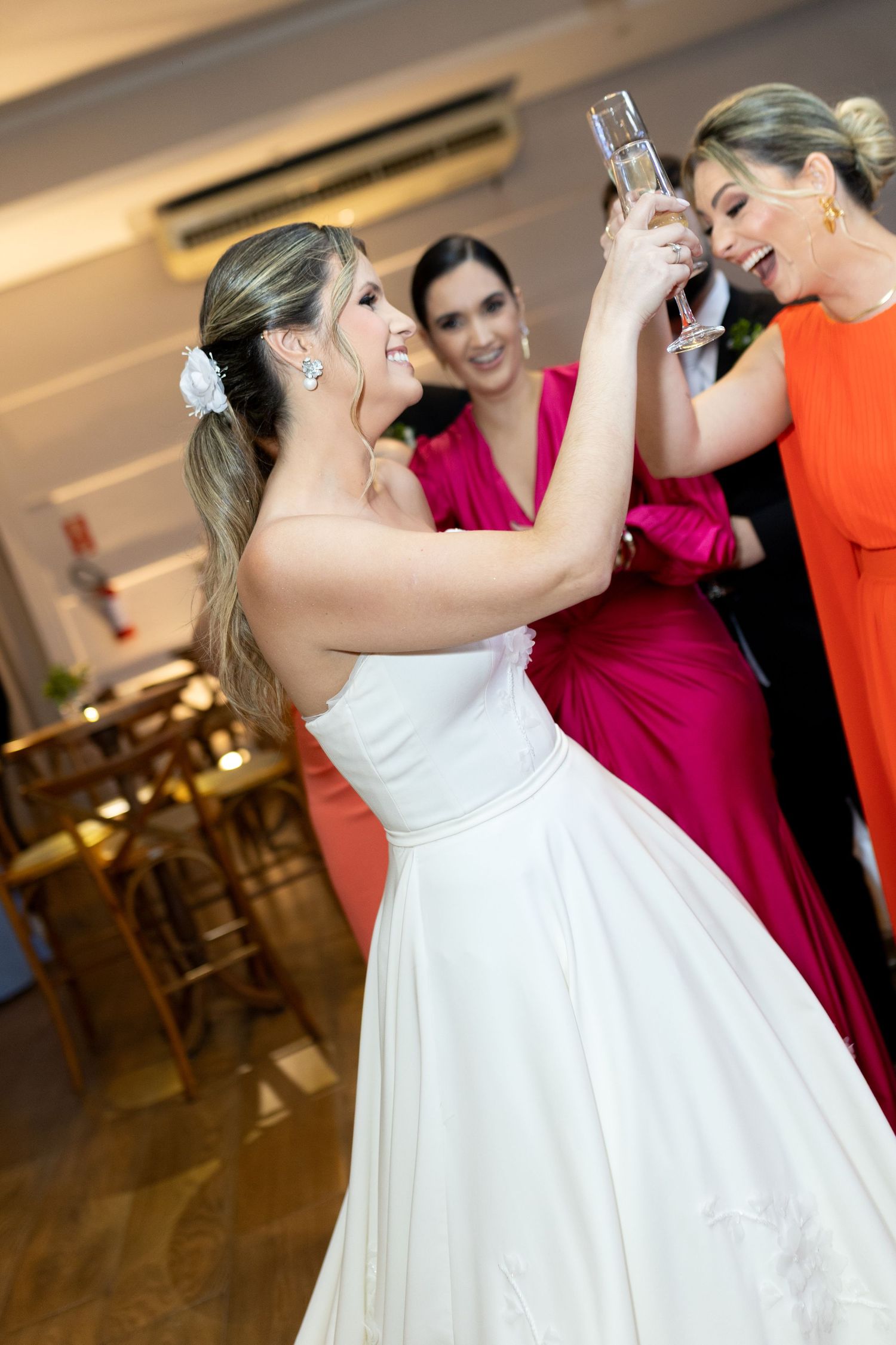 A smiling bride in a white gown toasts champagne with bridesmaids in pink and orange dresses at a wedding reception.