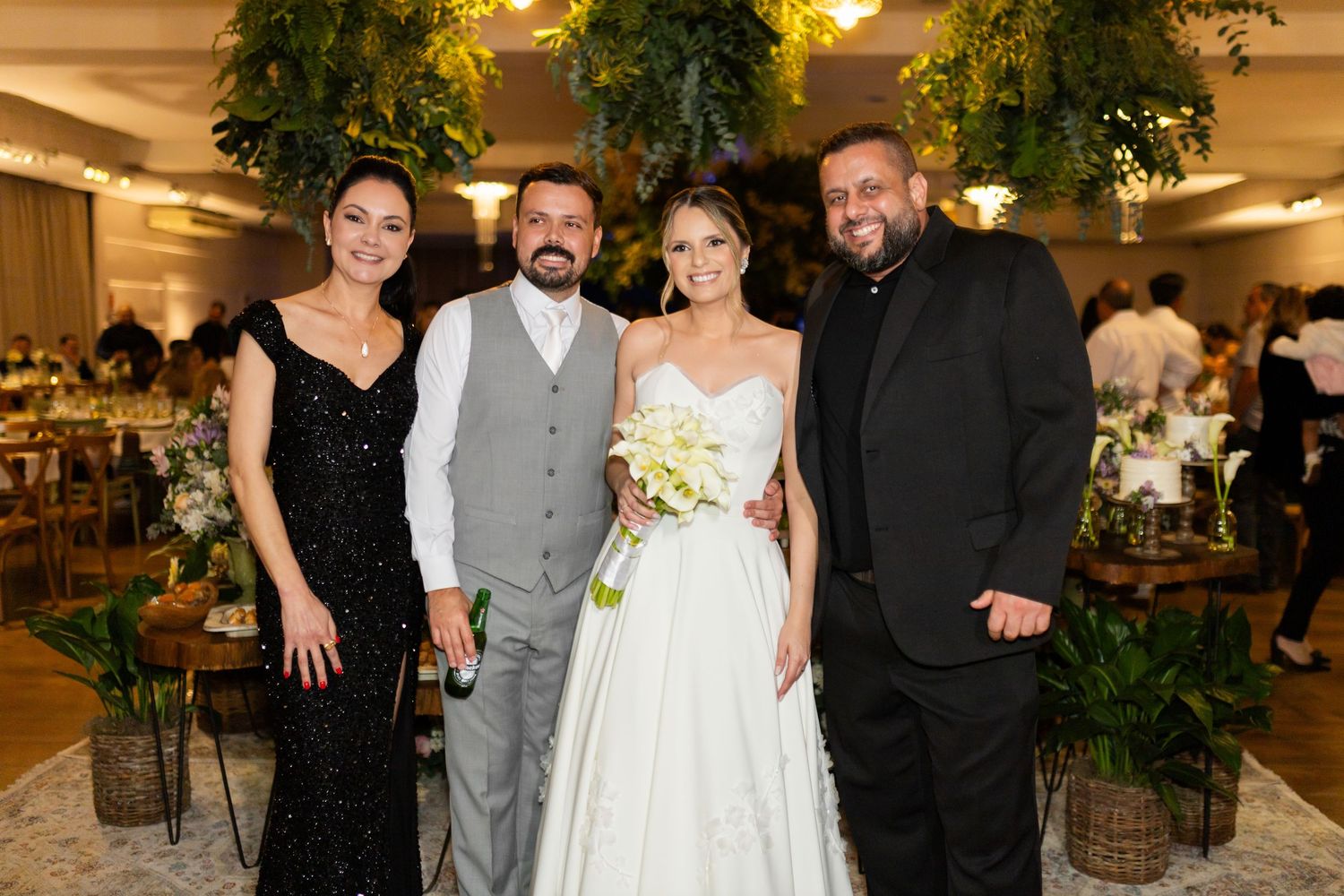 Bride and groom pose with two guests at elegant wedding reception with lush greenery decor.