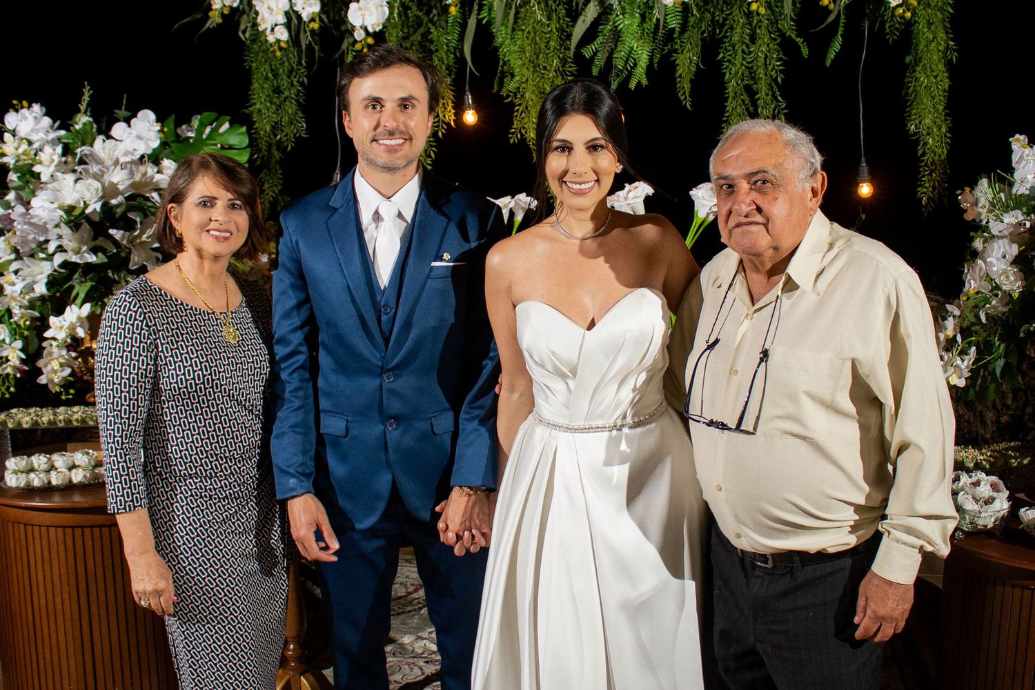 Bride and groom posing with elderly parents at elegant wedding reception with floral greenery backdrop.