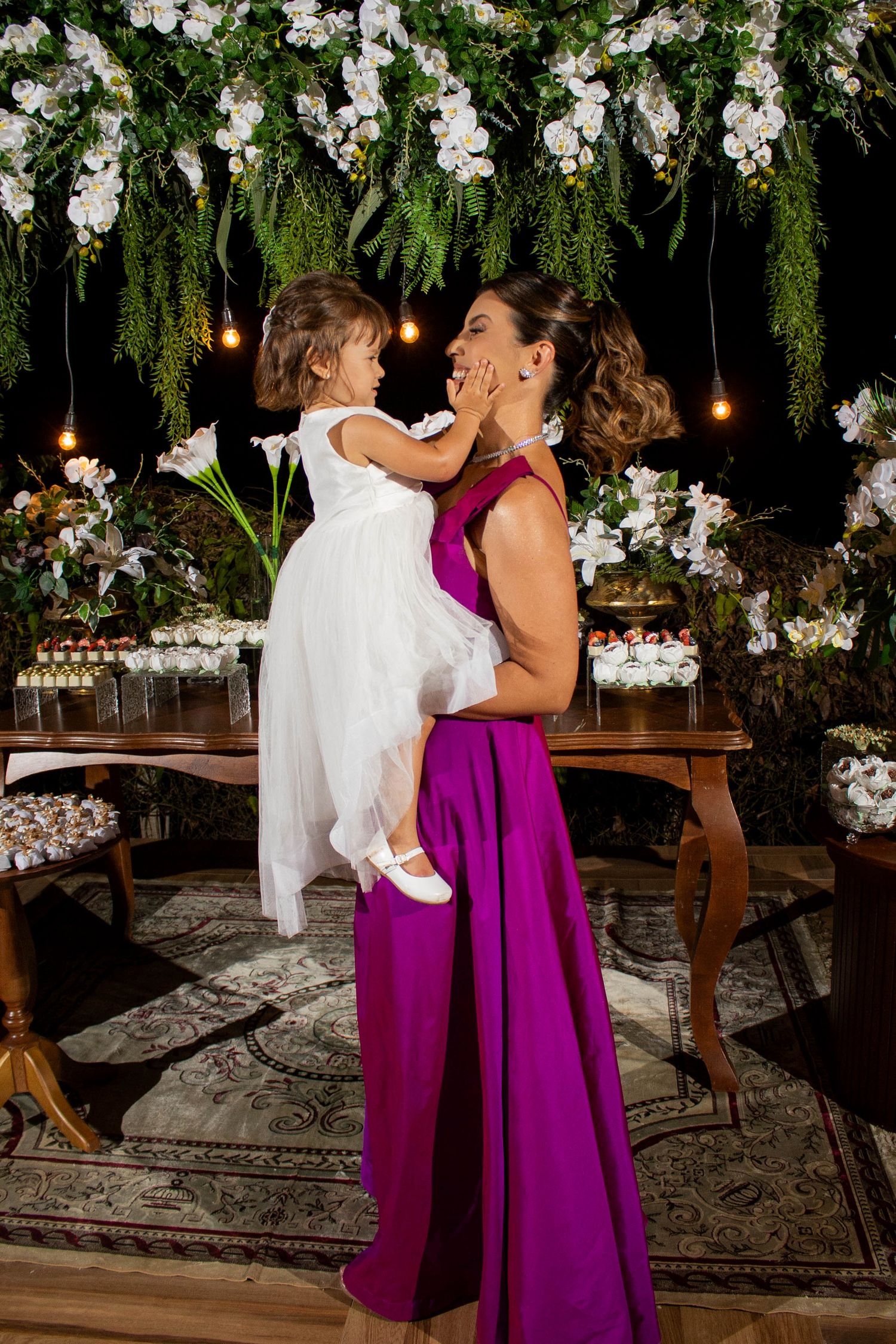 Woman in purple gown holds young girl in white dress at elegant floral event with greenery backdrop.