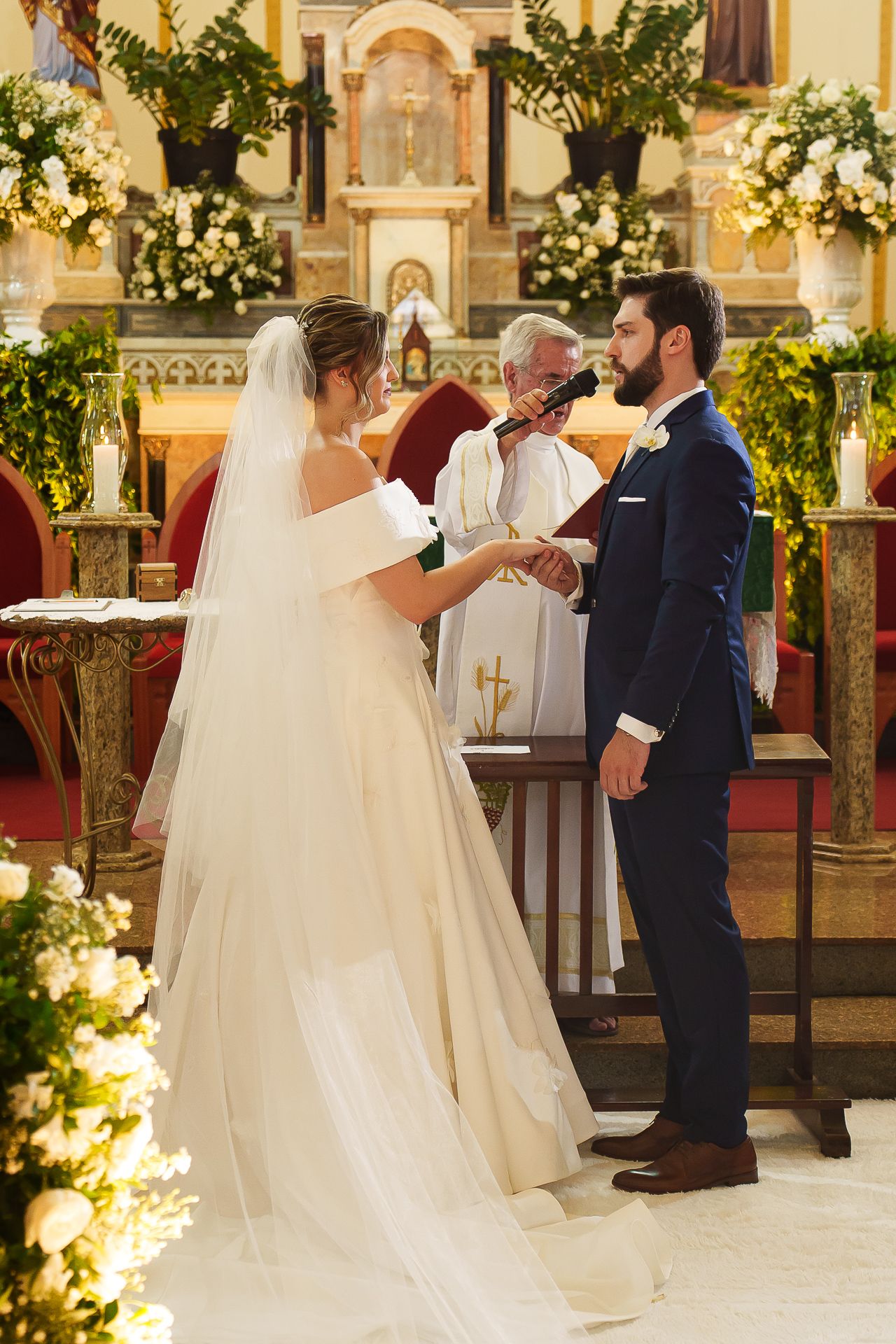 Bride and groom exchange vows at church altar, officiant behind them, surrounded by white floral arrangements.