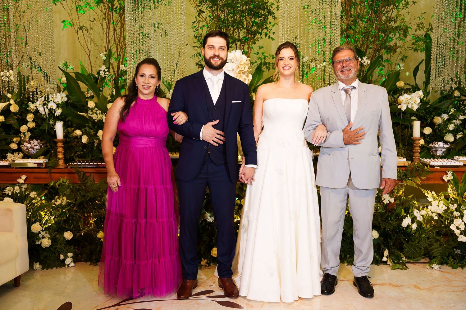 A bride and groom pose with family members at an elegant wedding reception with lush green floral backdrop.
