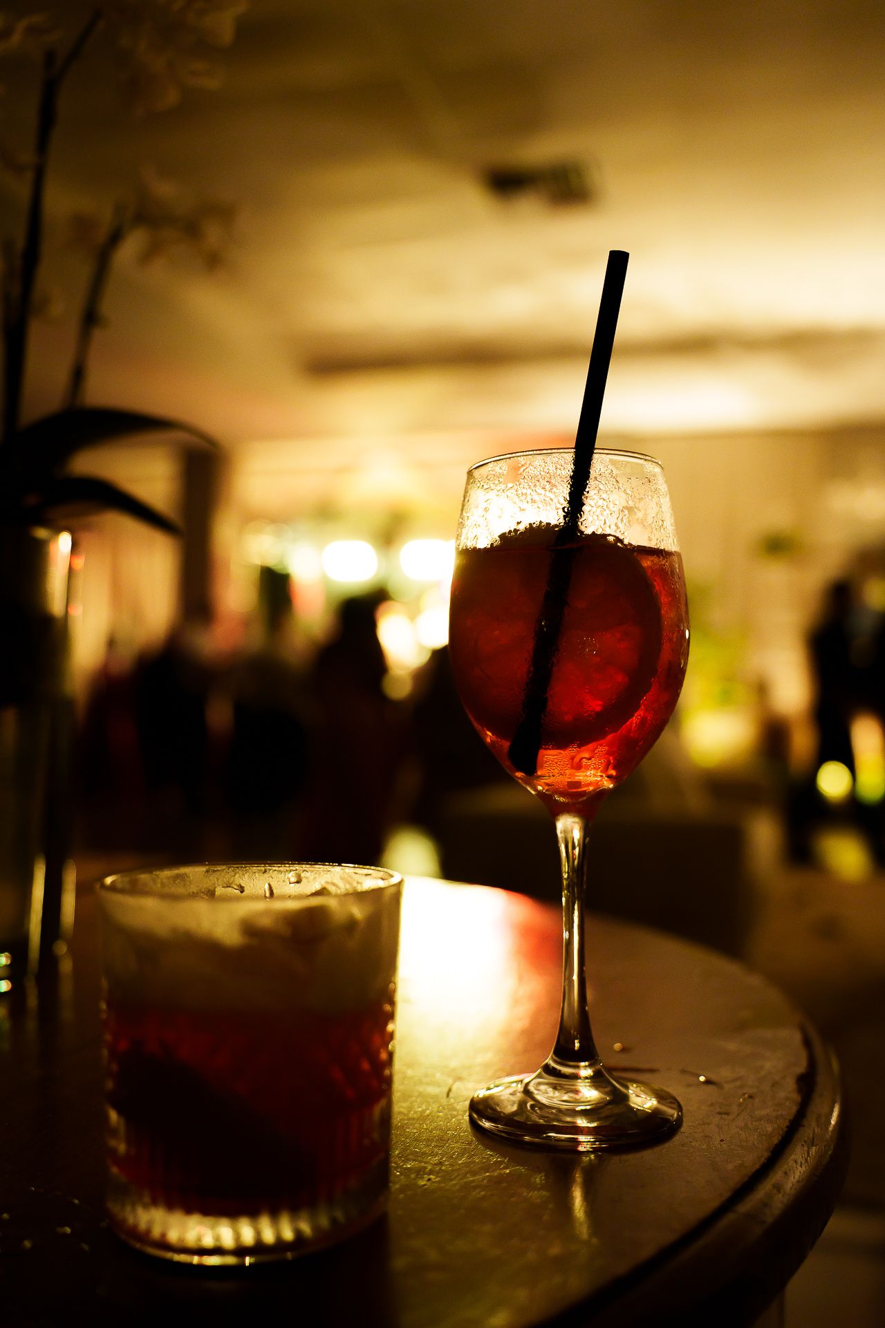 Two cocktails on a dark bar table, a wine glass with red drink and straw, and a rocks glass, in a dimly lit bar.