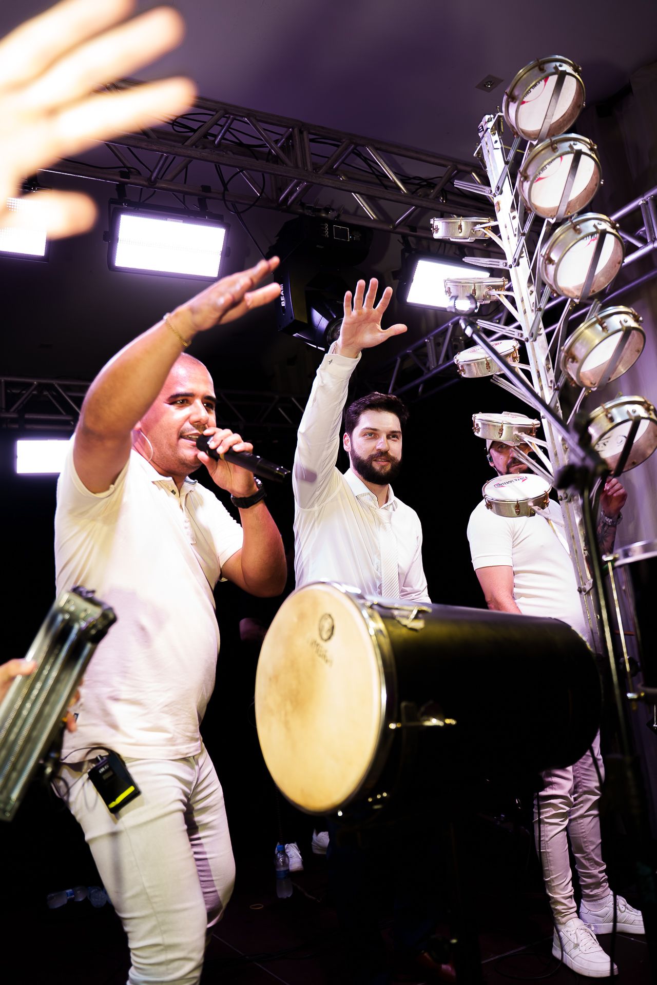 Three musicians in white perform on stage, with a vocalist singing into a microphone beside a large drum and decorative metal tree.