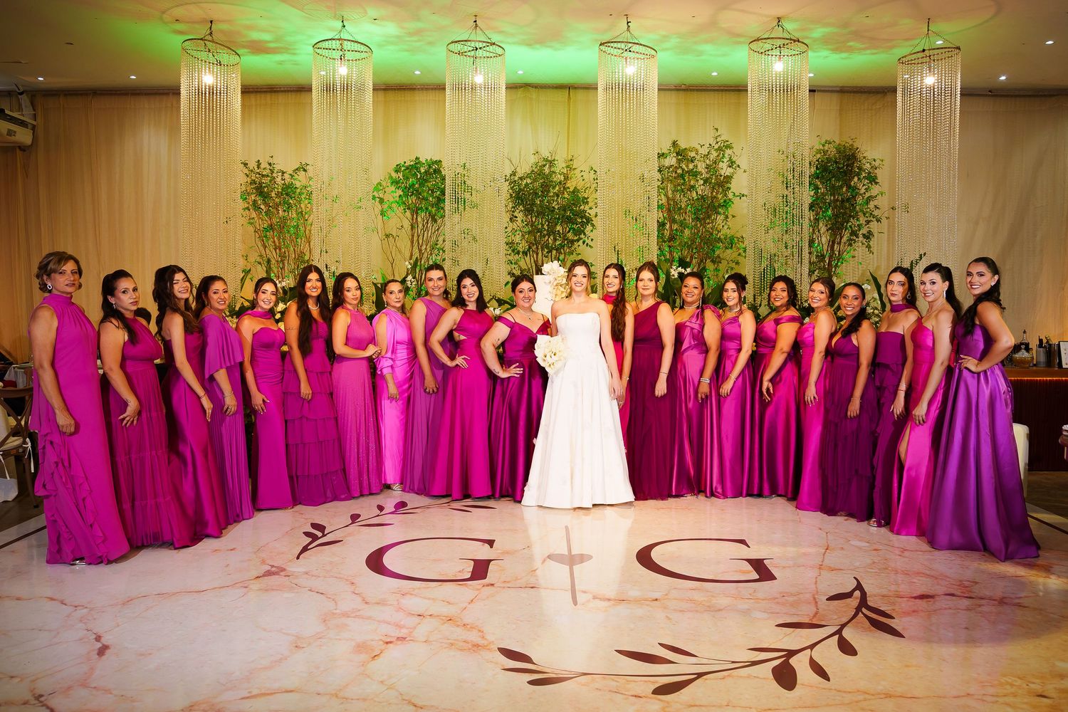 Bride in white gown poses with large bridal party in fuchsia gowns on monogrammed dance floor at elegant wedding reception.