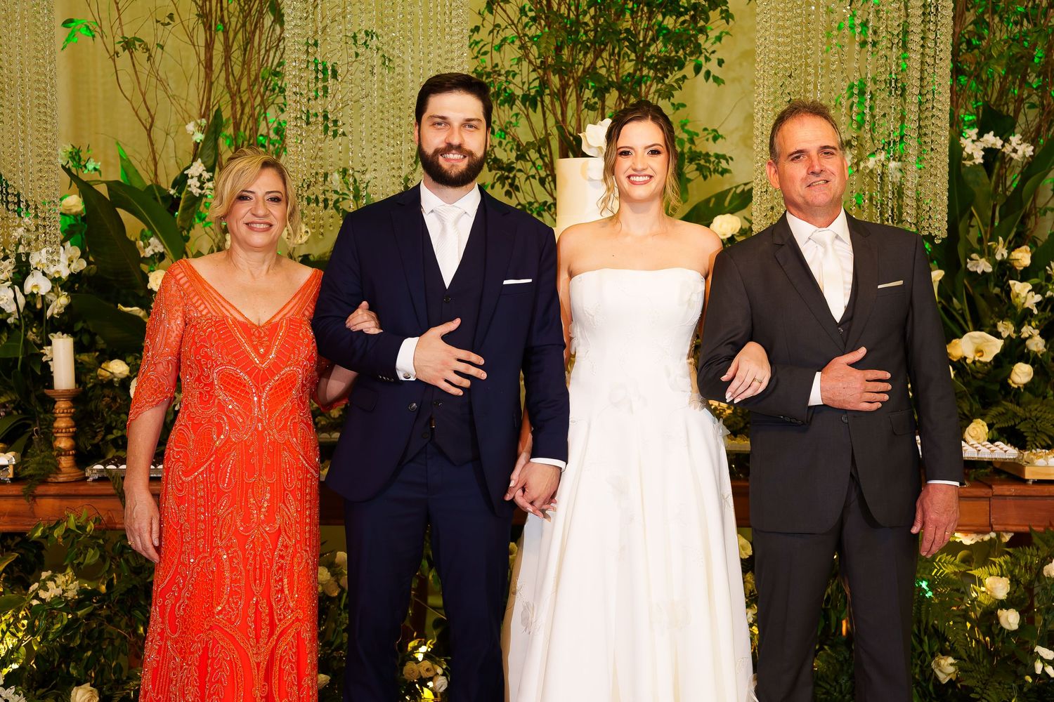 Wedding party poses together, bride in white gown, groom in navy suit, flanked by elegantly dressed family members.