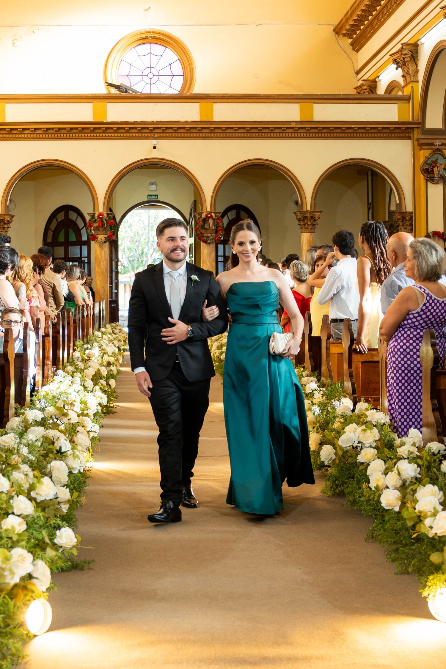 Couple walking down church aisle in teal gown and tuxedo, flanked by white floral arrangements.
