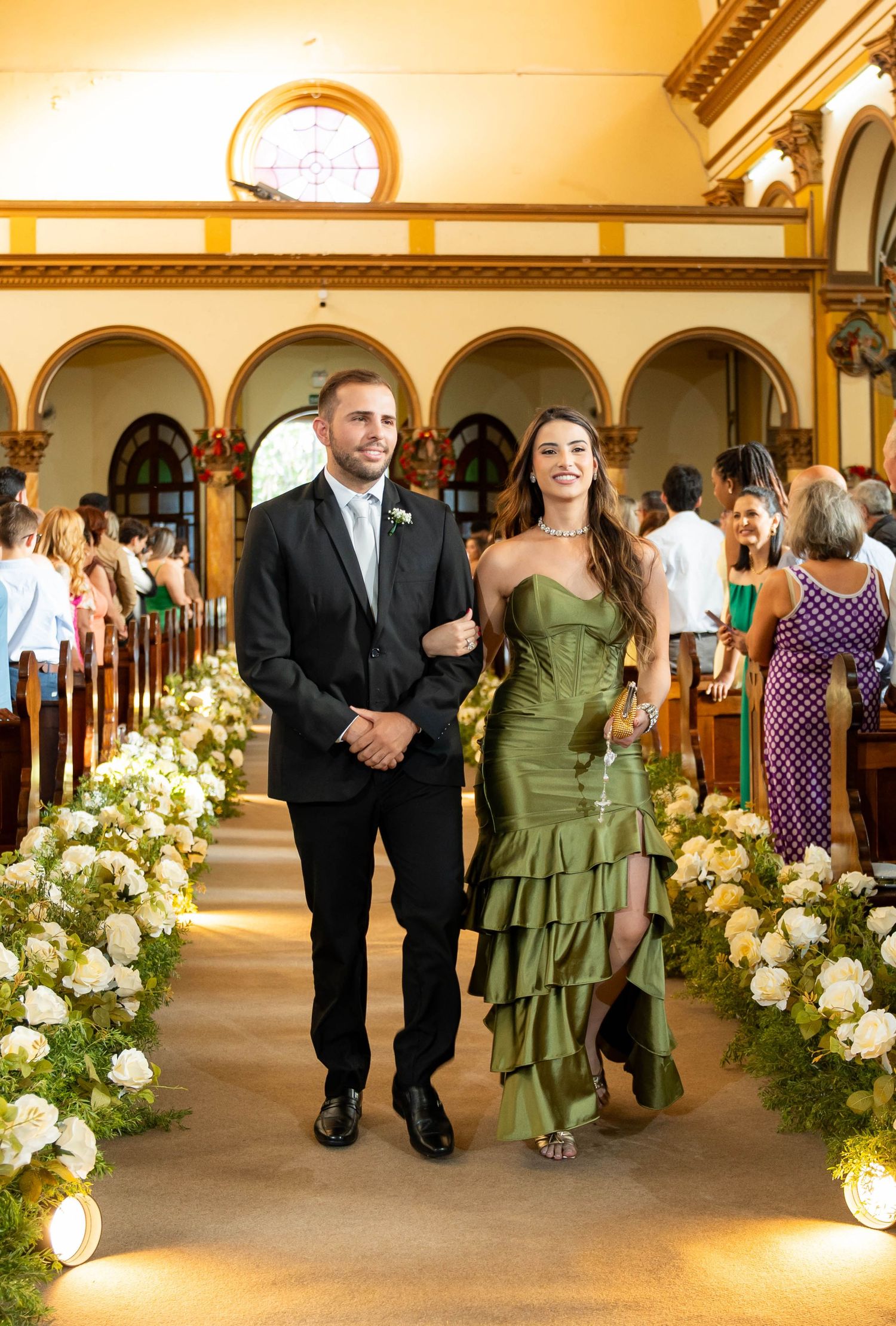 Couple walking down church aisle at wedding, man in black tuxedo and woman in green ruffled gown, flanked by white floral arrangements.