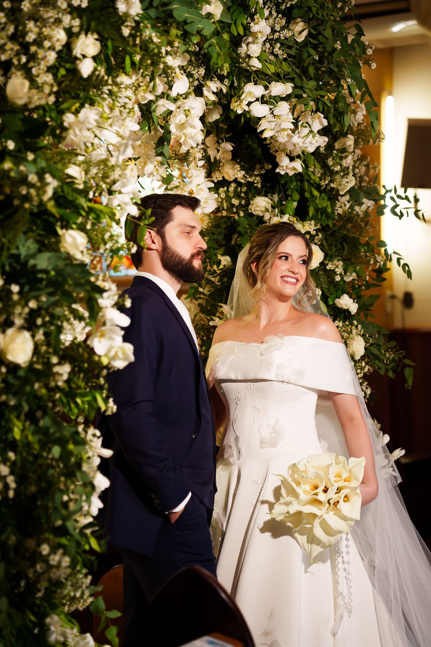 Bride in off-shoulder gown holding white bouquet stands with bearded groom in navy suit before floral arch.