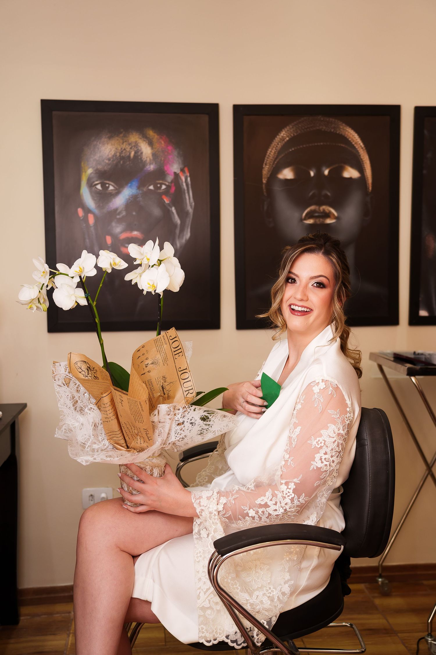 Smiling bride in white lace robe holding orchids sits in salon chair with colorful portrait art on wall behind her.