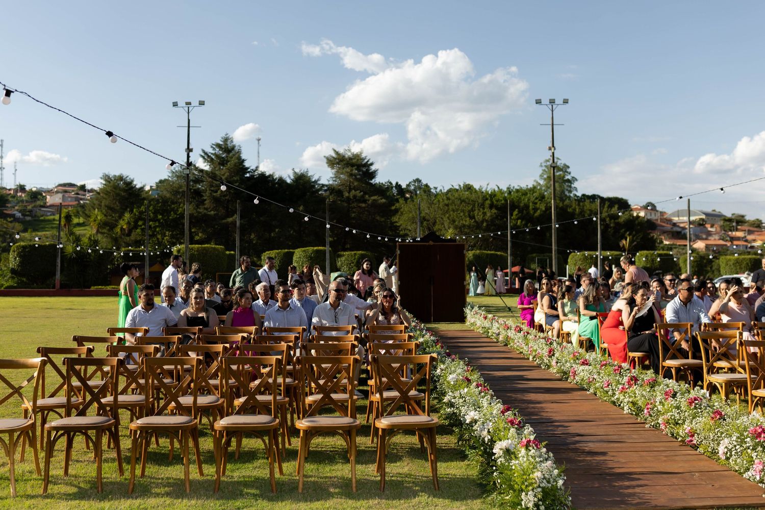 Outdoor wedding ceremony on green lawn with wooden chairs, floral aisle, string lights, and guests seated under blue sky.