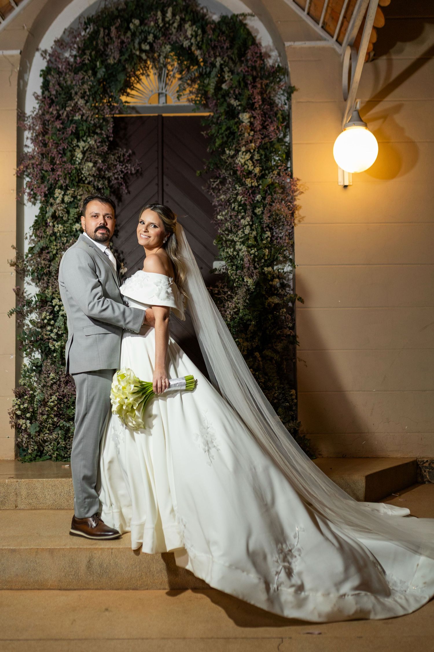 Bride and groom posing on church steps before floral arch doorway, bride in white ballgown with veil, groom in gray suit.