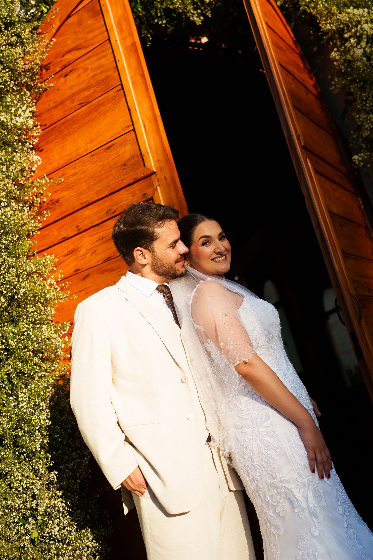 Bride and groom in white wedding attire pose romantically before rustic wooden chapel doors surrounded by lush greenery.