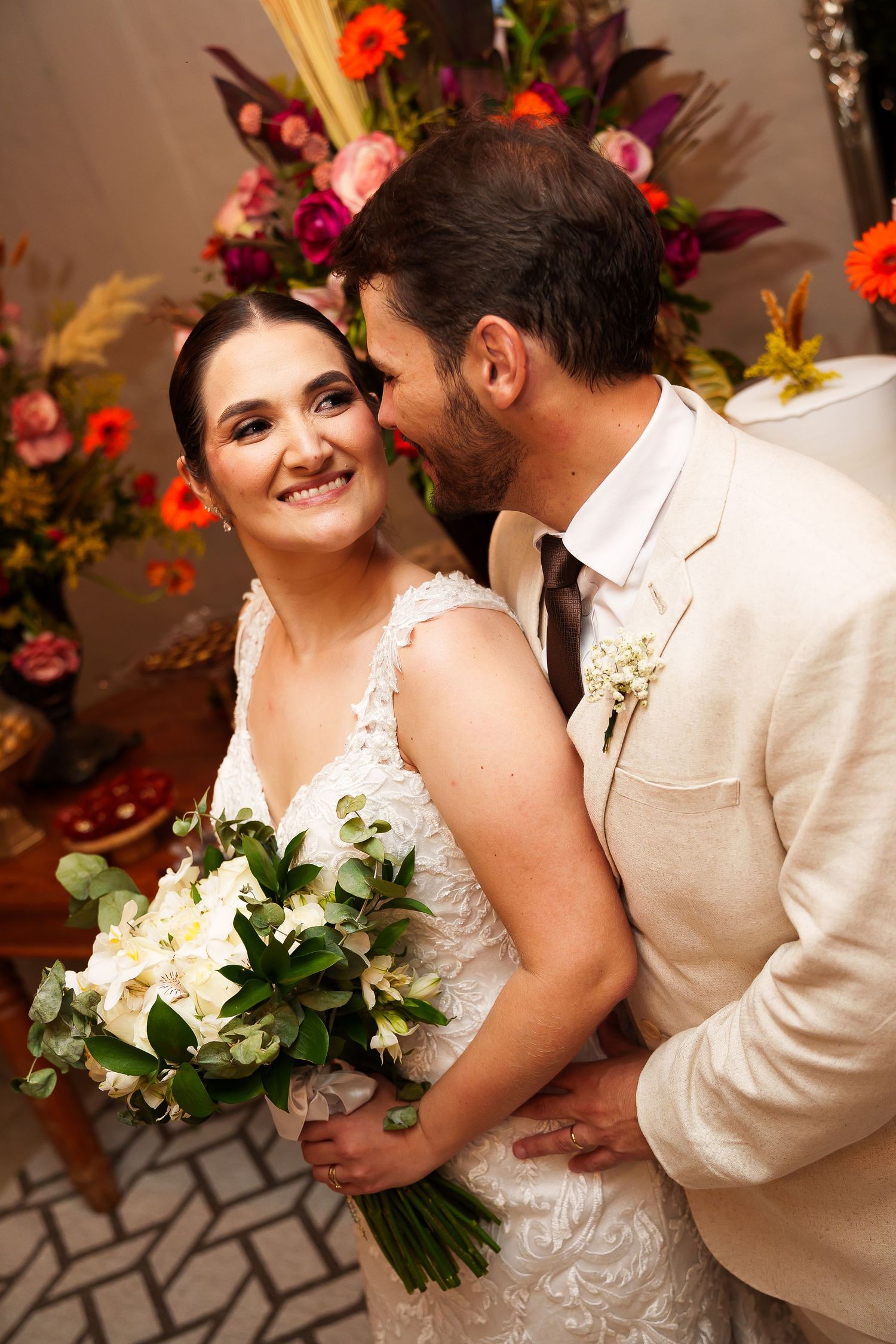 Bride smiling with bouquet as groom kisses her cheek at colorful floral wedding ceremony.