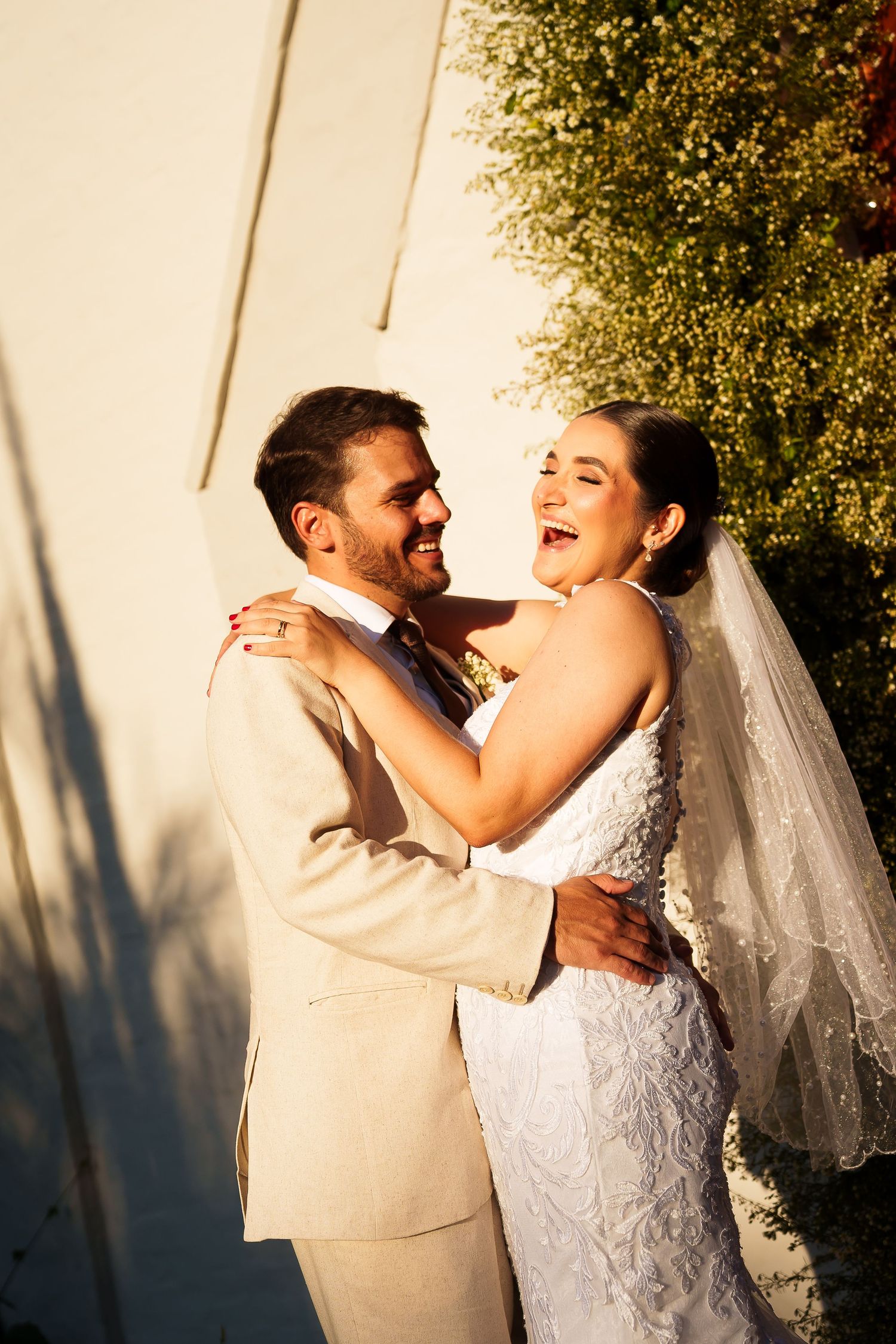 A joyful bride and groom embrace and laugh together outdoors in golden sunlight, she in a white gown and veil, he in a beige suit.