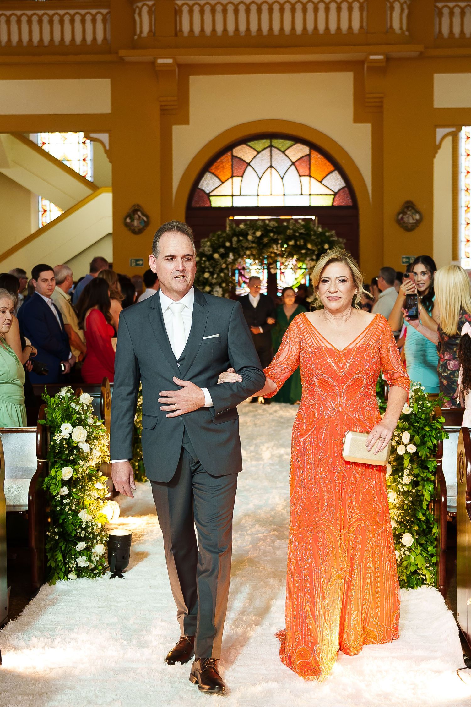 Couple walking down church wedding aisle, man in gray suit and woman in orange sequin gown, floral decorations.
