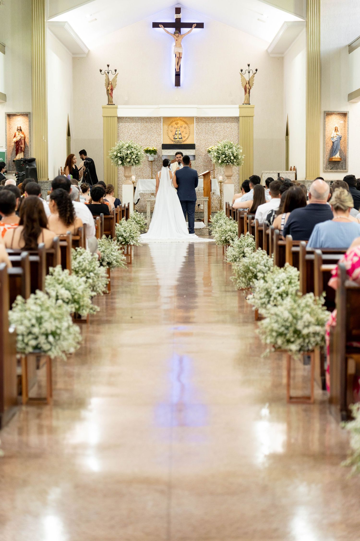 A couple exchanges vows at the altar of a Catholic church, with white floral arrangements lining the aisle and guests seated in wooden pews.